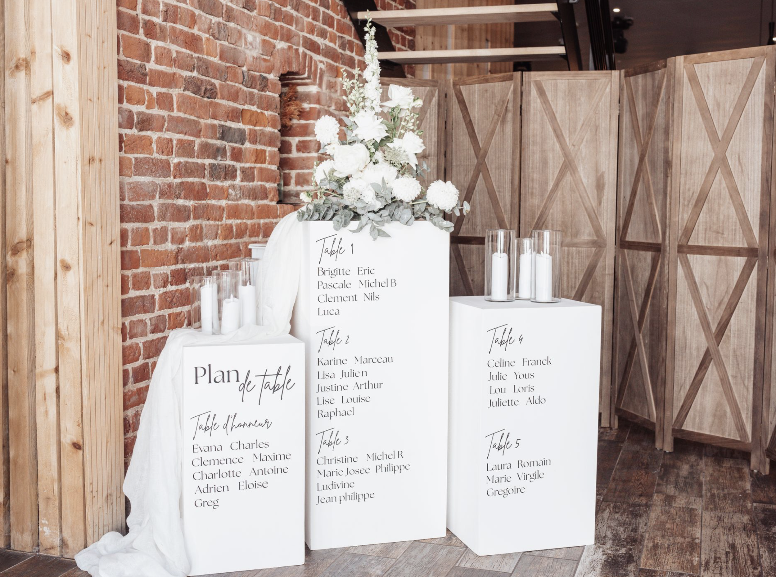 Tableau de mariage avec une décoration florale blanche et verte, plan de table écrit à la main sur des panneaux blancs, entouré de chandelles dans des verres, fond en briques rouges et panneaux en bois
