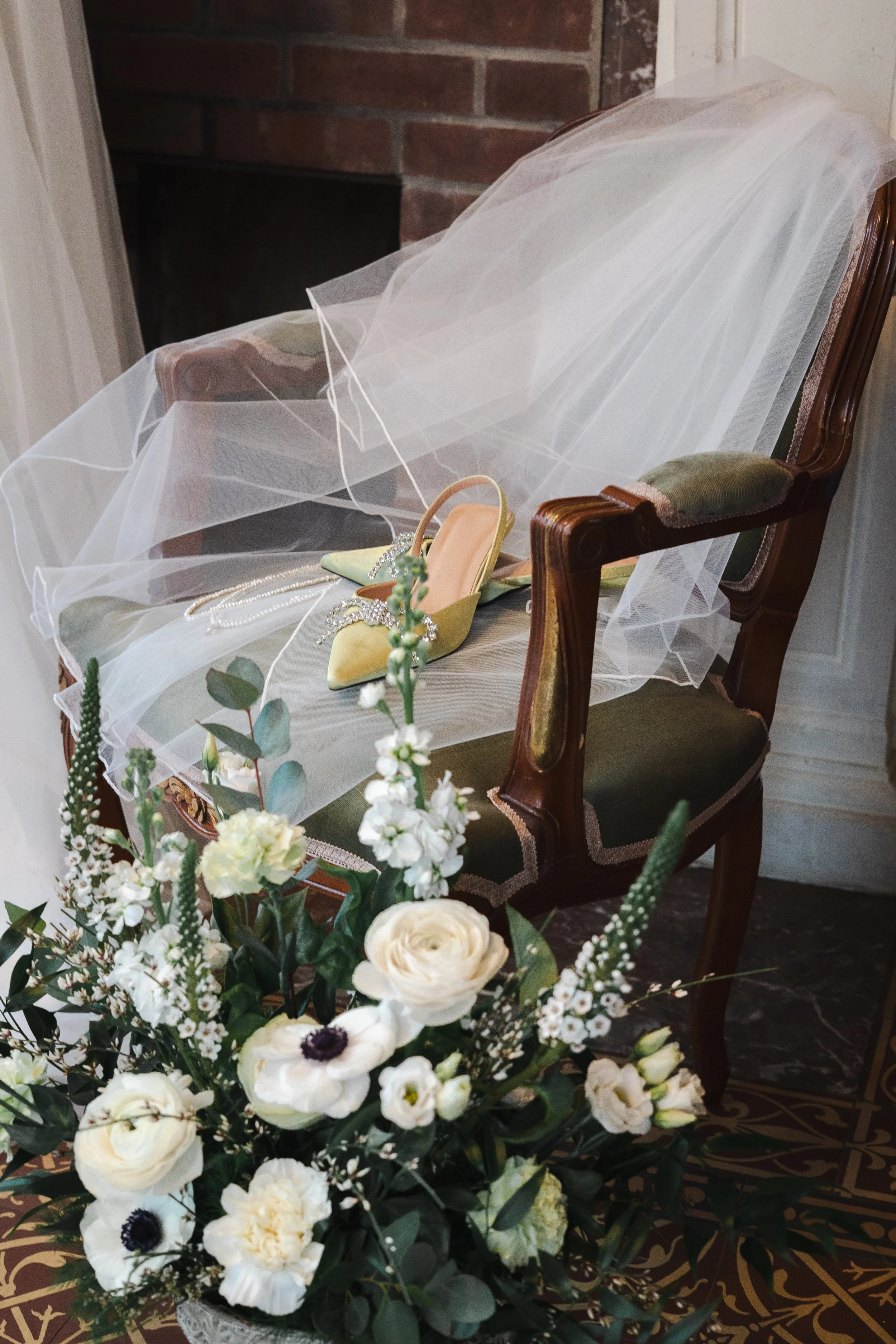 Une chaise ancienne en bois avec un voilage blanc, une paire de chaussures à talons vert clair et des bijoux posés dessus, et un bouquet de fleurs blanches en premier plan.