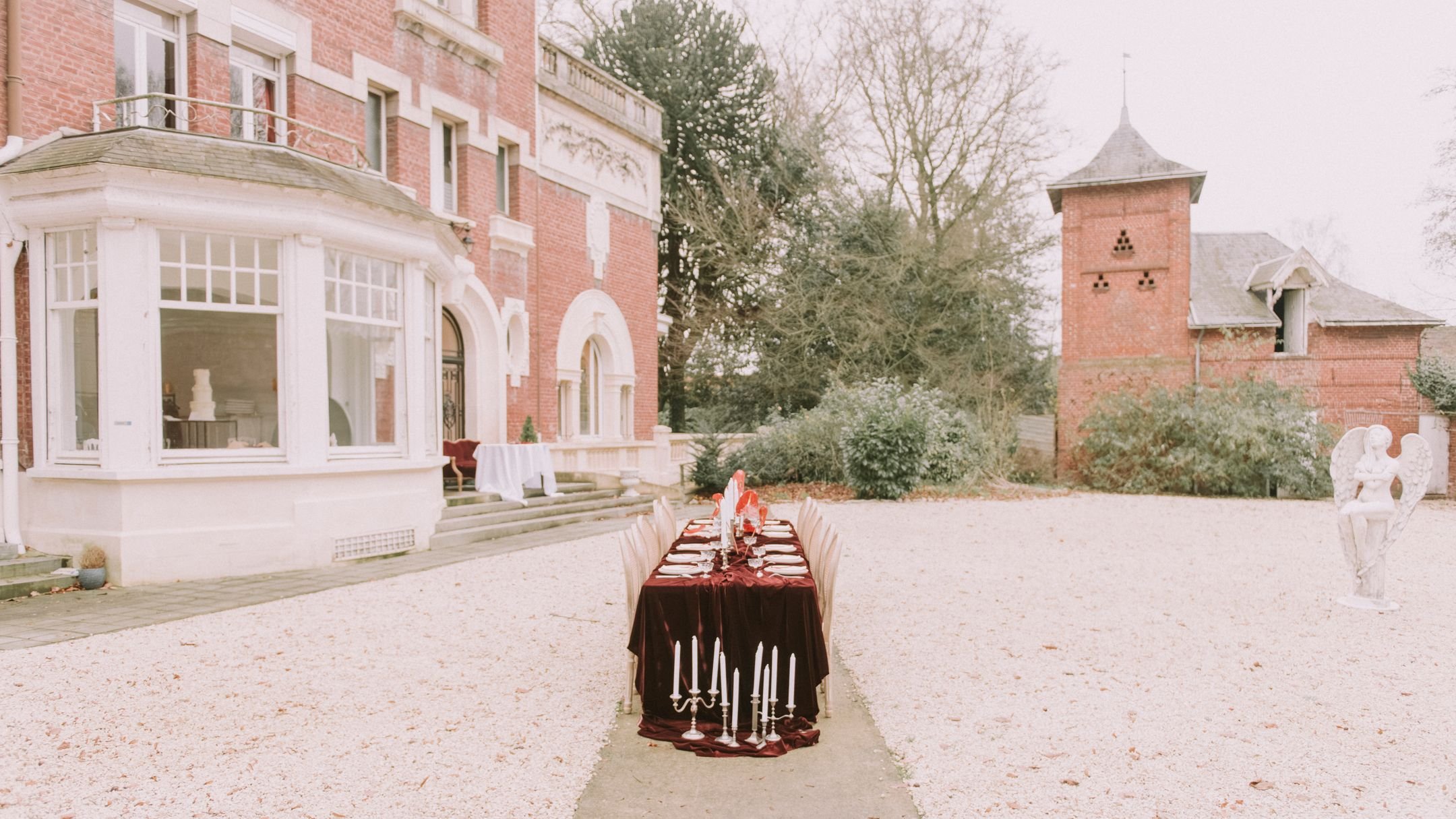 Une table de banquet avec une nappe rouge foncé, des chandeliers et des plats, dressée dans une cour extérieure d'un bâtiment ancien en briques rouges, avec une statue d'ange et un bâtiment avec une tour en arrière-plan.