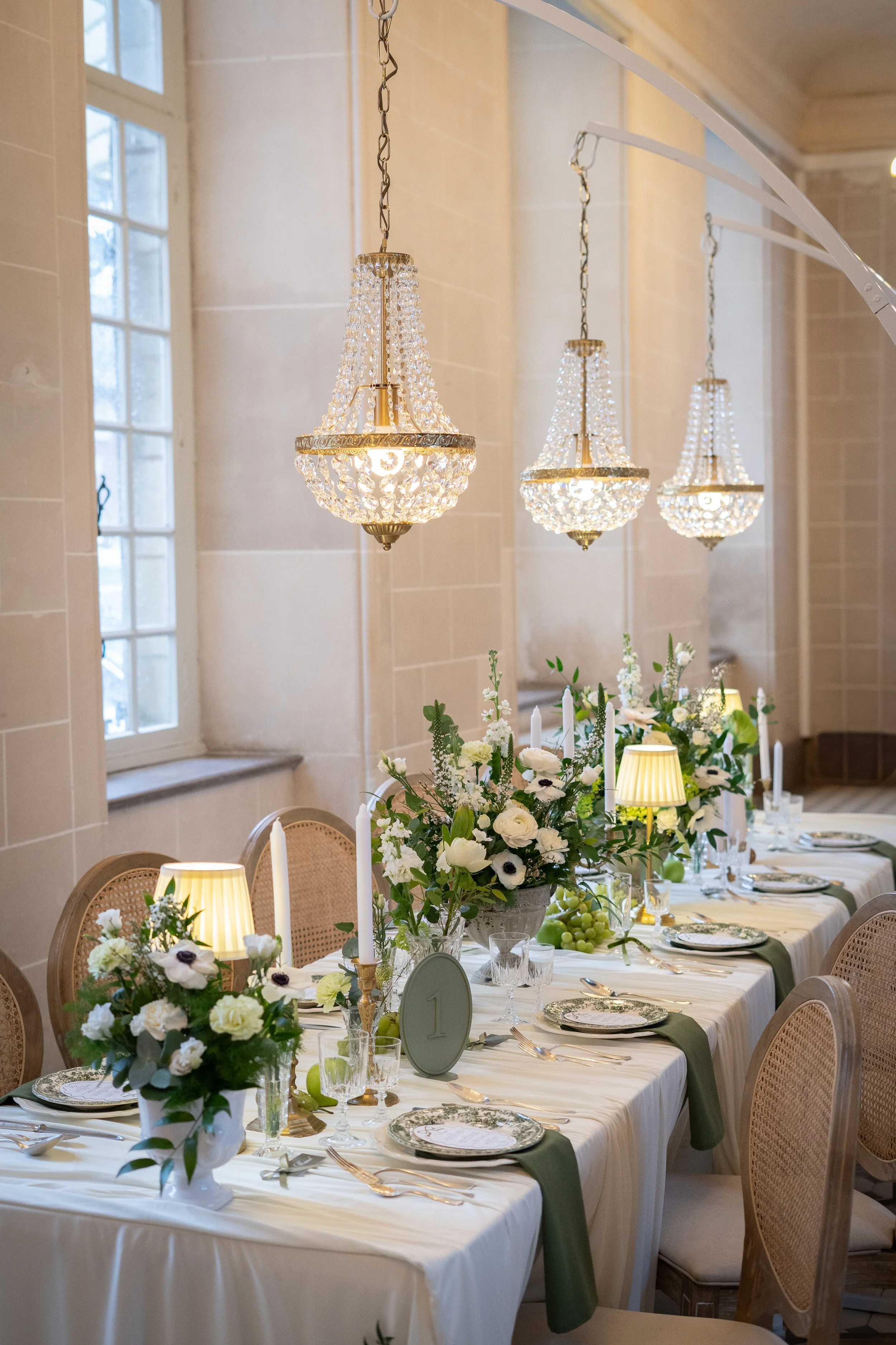 Salle de dîner élégante avec chandeliers en cristal, fleurs blanches, bougies, table dressée pour un repas, et lampes d'appoint.