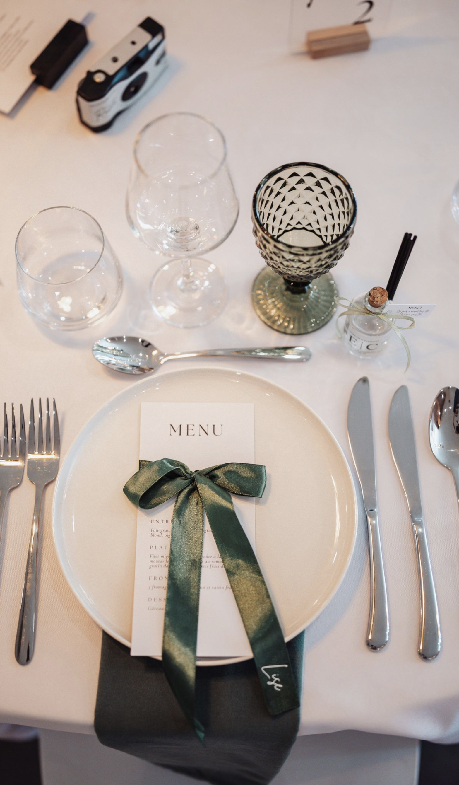 Table élégante préparée pour un repas, avec une assiette blanche, un menu attaché avec un ruban vert, un couteau et une fourchette en acier inoxydable, des verres à vin, un verre à eau, une petite bouteille avec un ruban et une étiquette, et un fond 