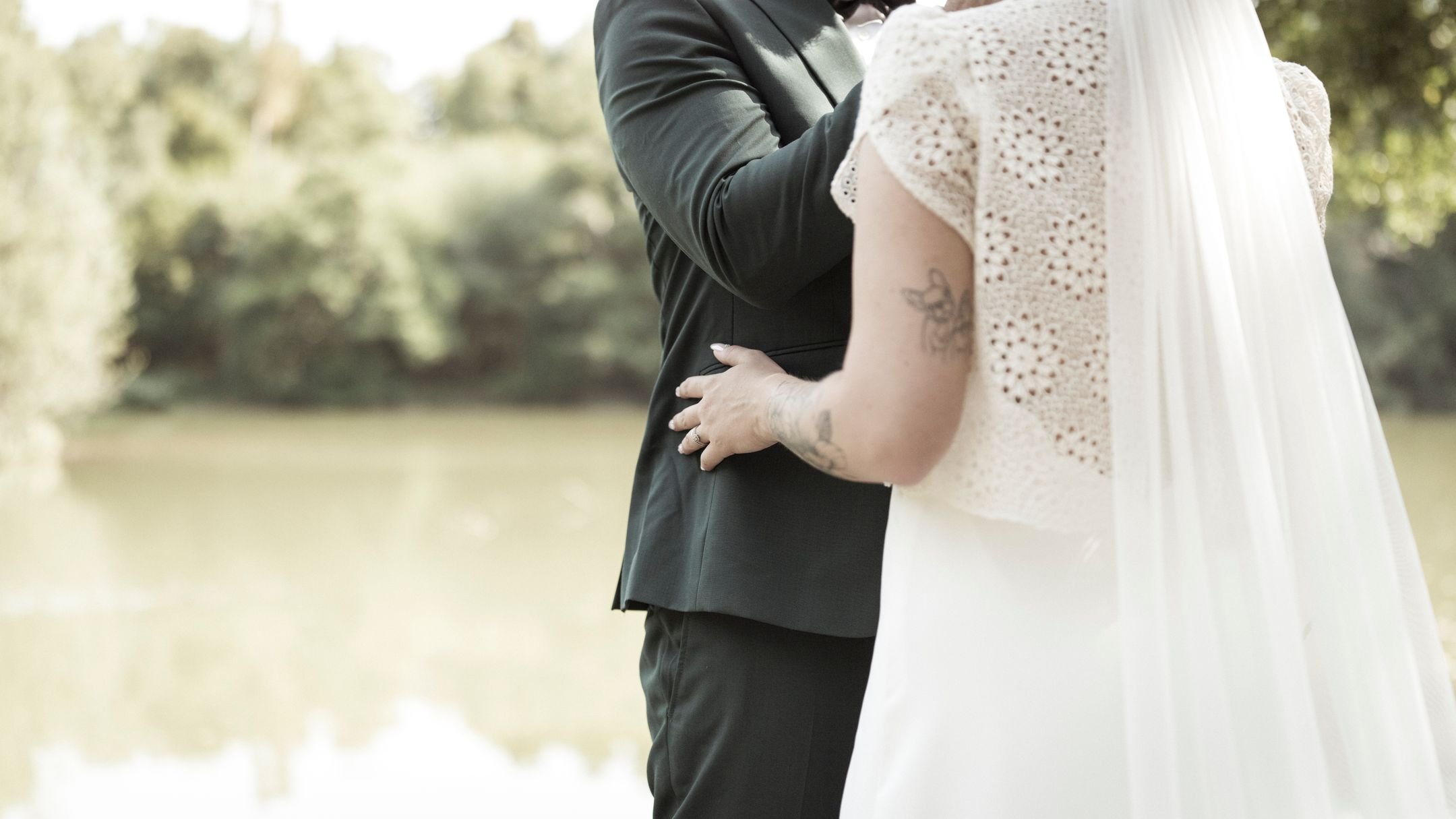 Un couple marié se tient près d'un plan d'eau lors d'une cérémonie de mariage en plein air, avec des arbres en arrière-plan.