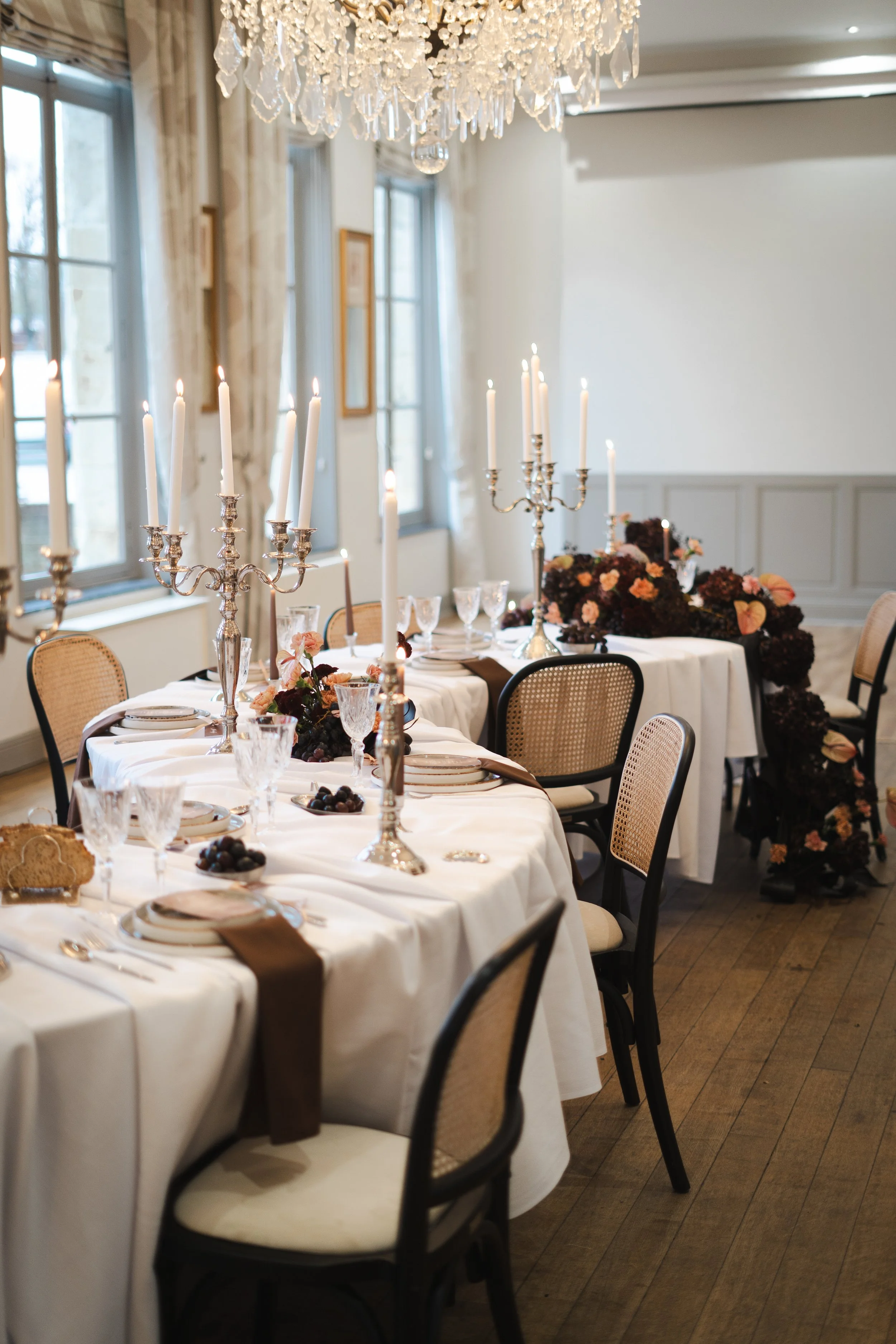 Salle à manger élégante avec table dressée, chandeliers en argent avec bougies allumées, décor floral de couleurs sombres et meubles en bois