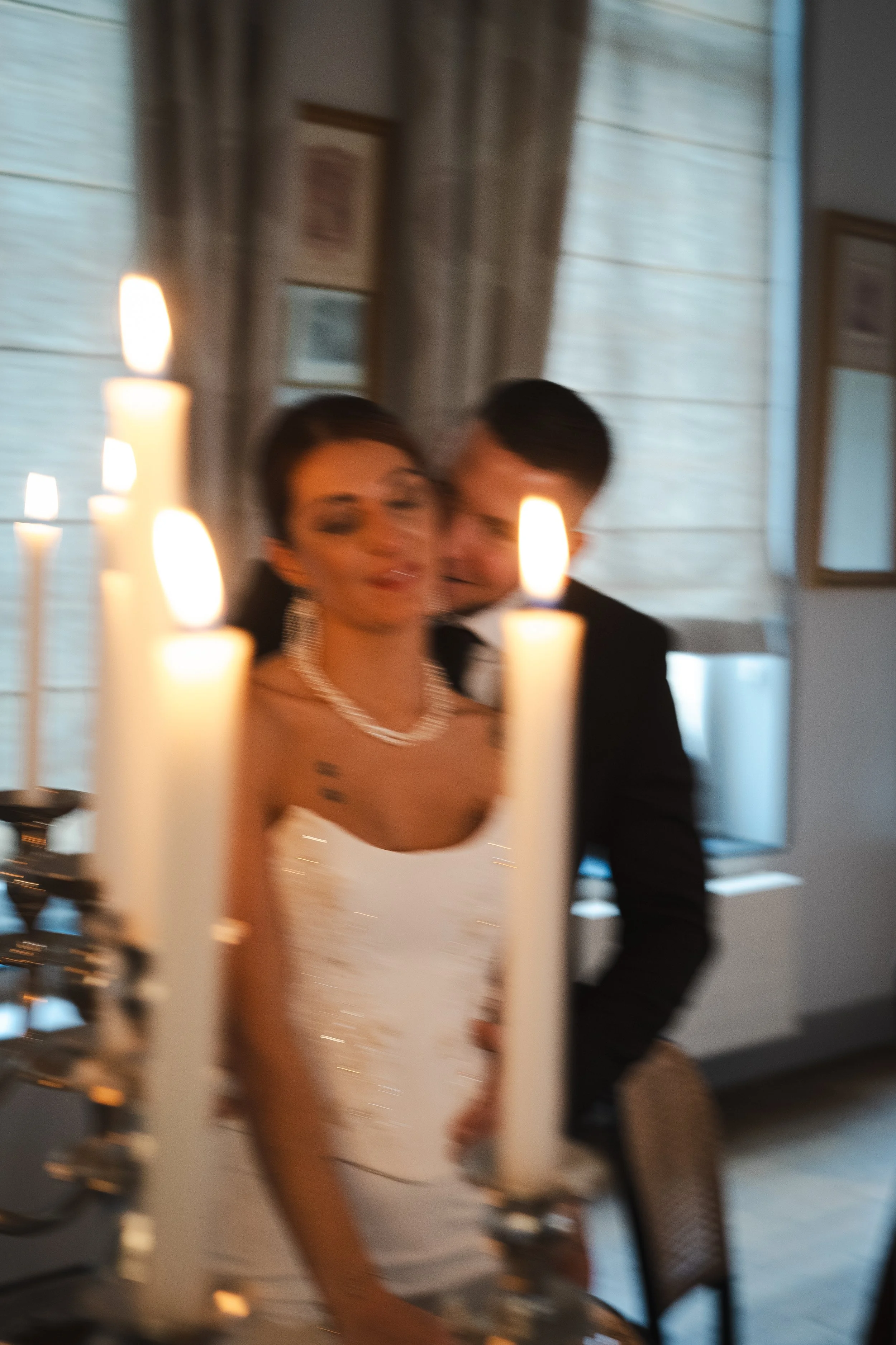 Deux personnes habillées formellement, une femme en robe blanche et un homme en costume noir, se tenant près d'une cheminée à bougies, dans une ambiance chaleureuse. La photo est floue.