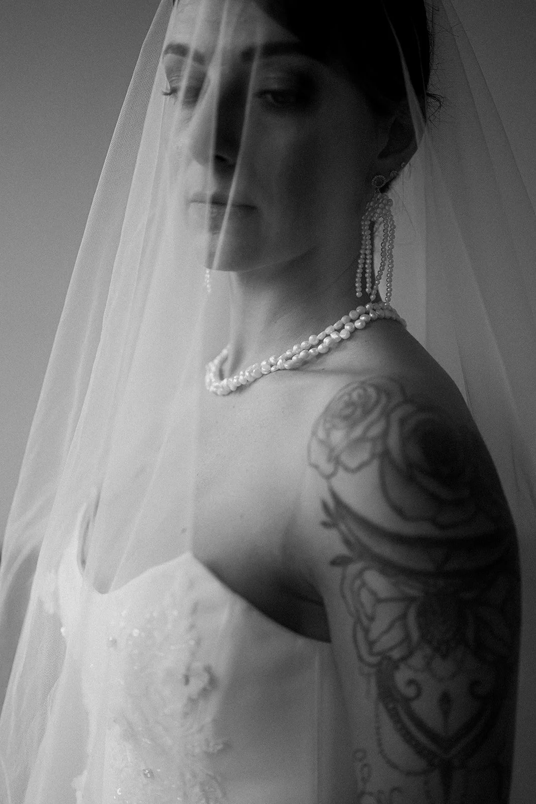 Une femme en robe de mariée avec un voile, portant des bijoux en perles et une tatouage sur le bras, regardant vers le bas en noir et blanc.
