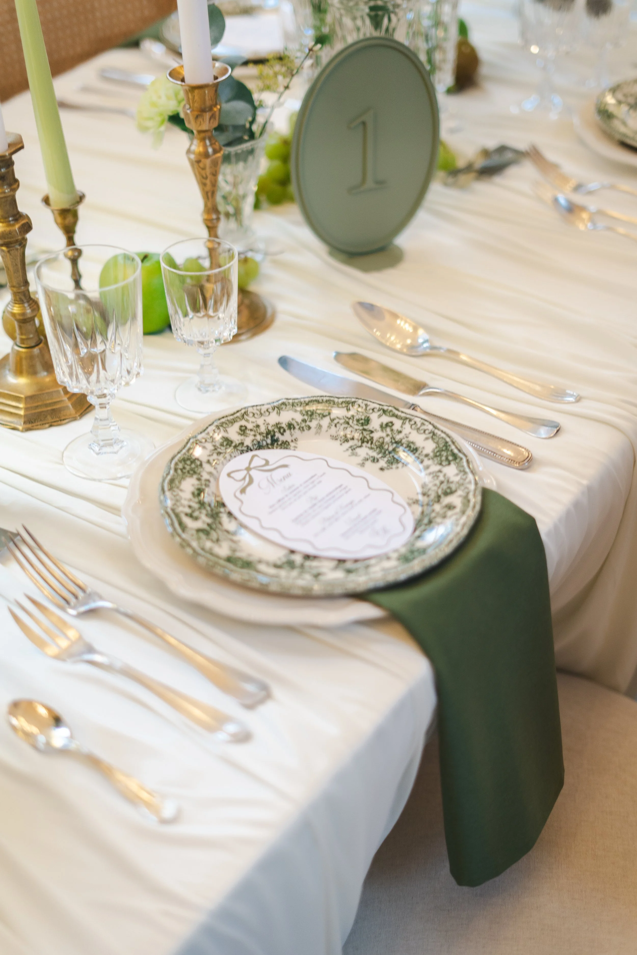 Table de mariage ou de banquet avec une nappe blanche, une assiette décorative verte et blanche, des couverts en argent, des verres à vin, des chandeliers en laiton avec des bougies vertes et blanches, un menu placé sur l'assiette, une petite table ronde verte avec le chiffre 1, et des décorations florales avec des feuilles vertes et des pommes vertes.