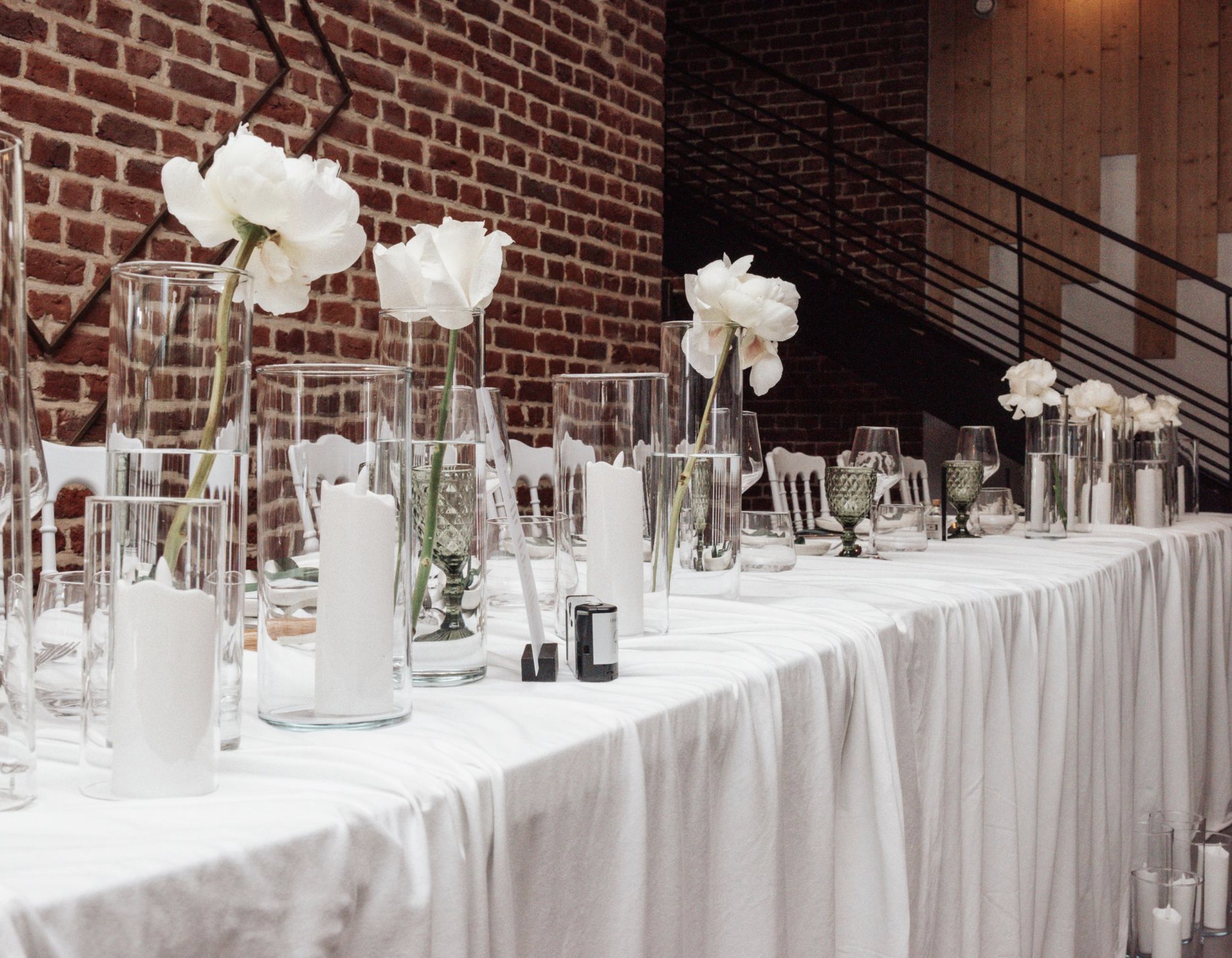 Une longue table décorée avec des vases contenant des fleurs blanches, des bougies et des verres à vin, dans une salle avec un mur en briques apparentes et une mezzanine en bois.