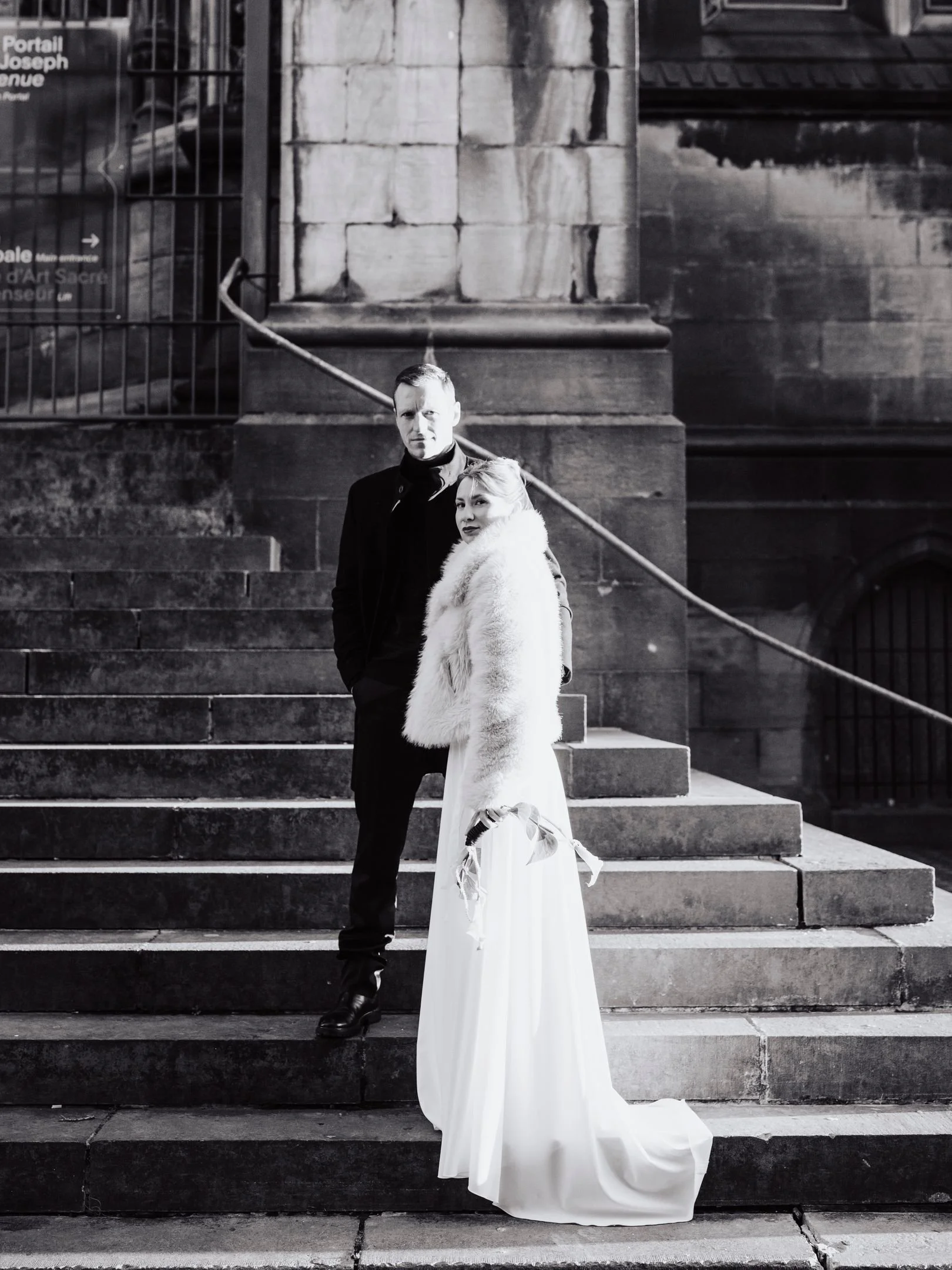 Deux personnes habillées élégamment, un homme en costume noir et une femme en robe blanche avec une fausse fourrure blanche, posant sur des escaliers en pierre devant un bâtiment en pierre, dans une scène en noir et blanc.