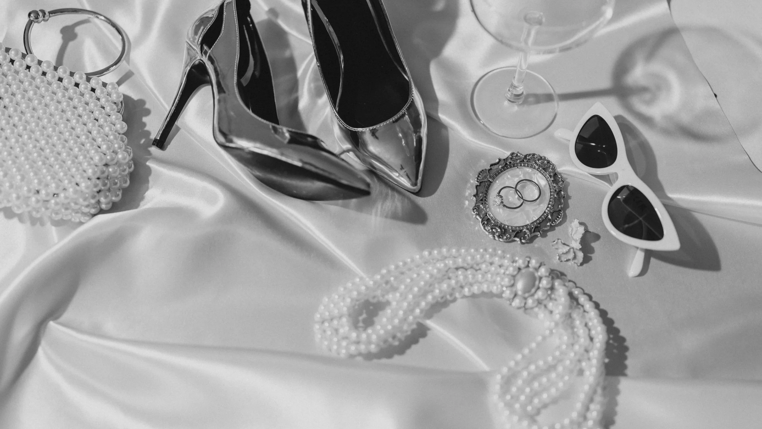 Accessoires de mode pour une occasion spéciale, comprenant des chaussures à talons en miroir, une pochette en perles, des lunettes de soleil blanches à cœur, des boucles d'oreilles et un bracelet en perles, sur une surface en satin.