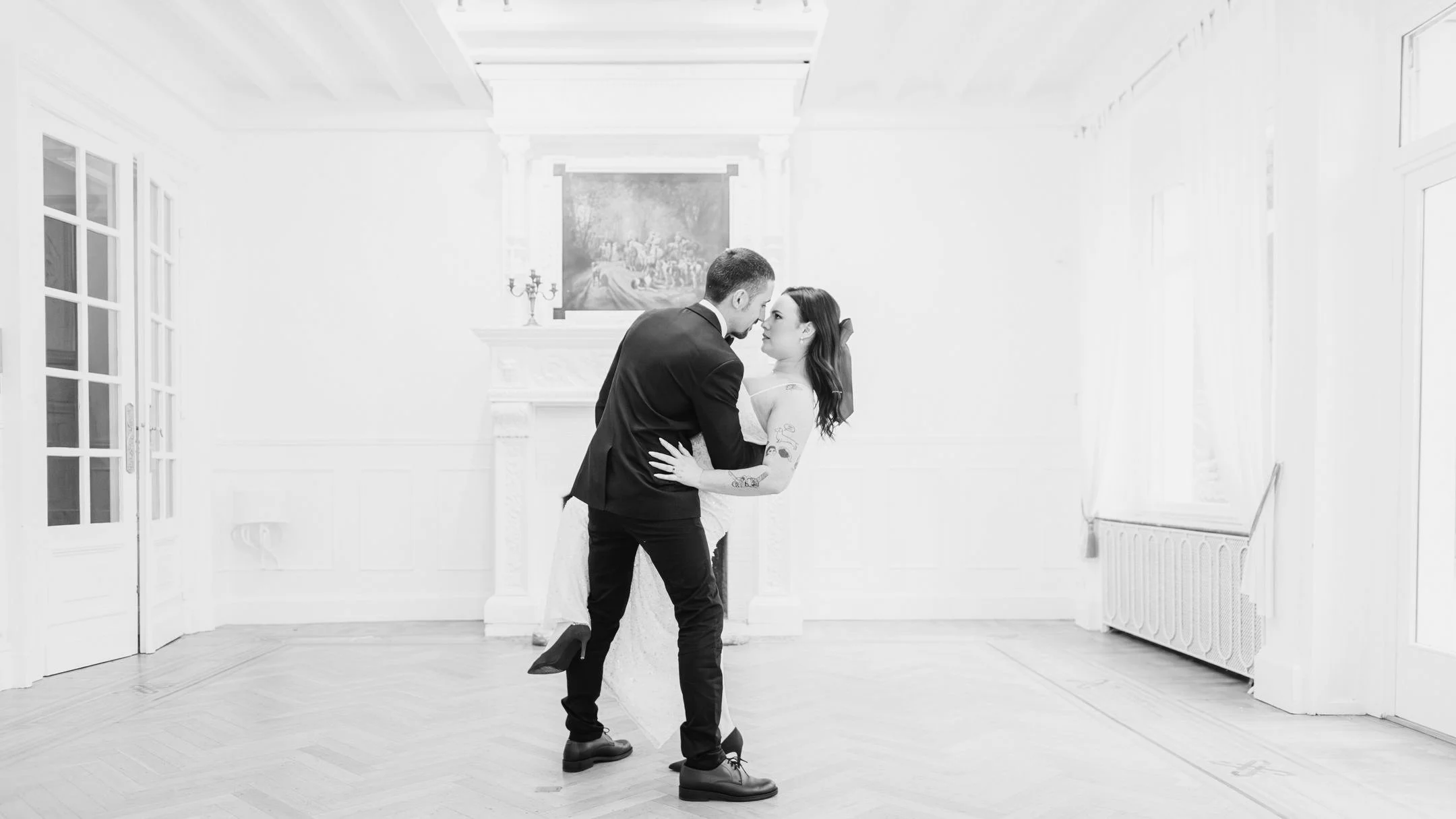 Un couple danse dans une pièce lumineuse avec un décor classique, murs blancs, cheminée et peinture au mur.