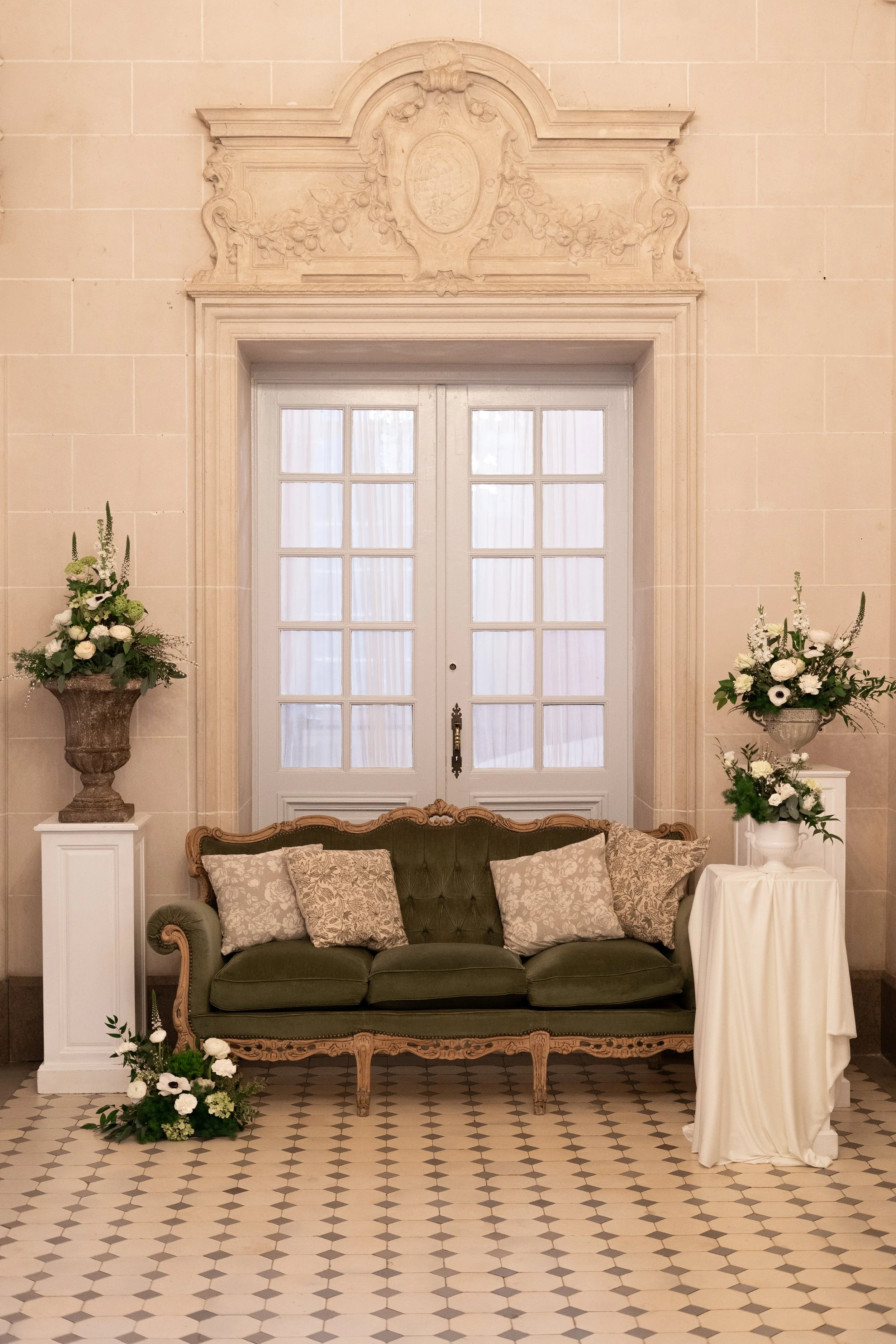 Un fauteuil ancien vert avec des coussins beige, flanqué de deux grands vases floraux blancs avec des fleurs blanches et vertes, devant une porte en verre avec rideaux blancs, dans une pièce élégante.