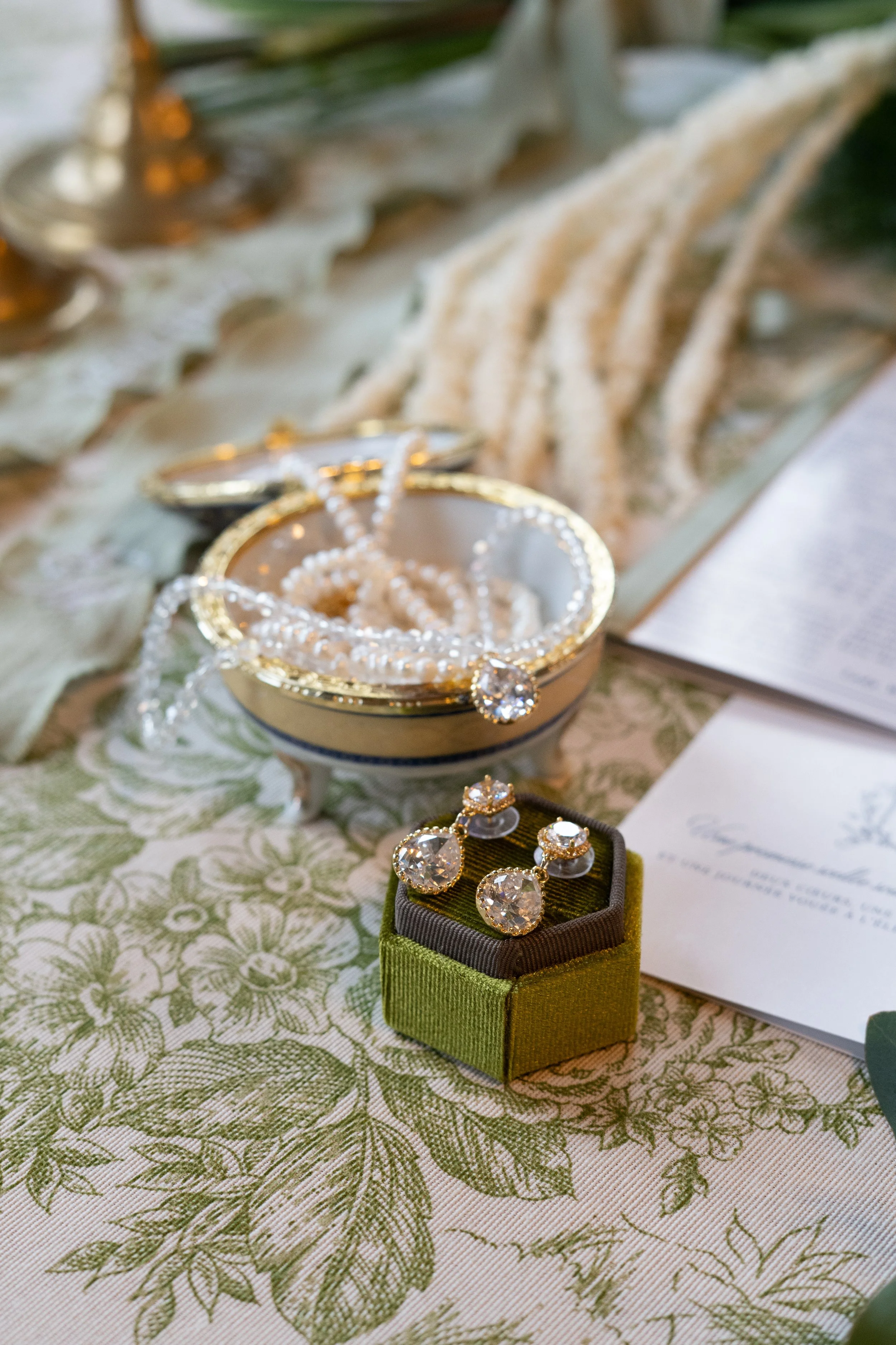 Bijoux en diamants, y compris une bague, des boucles d'oreilles, et un pendentif présentés sur une boîte en velours vert. Autour, un bol contenant des perles de perle, un napperon de table floral, et des papiers.