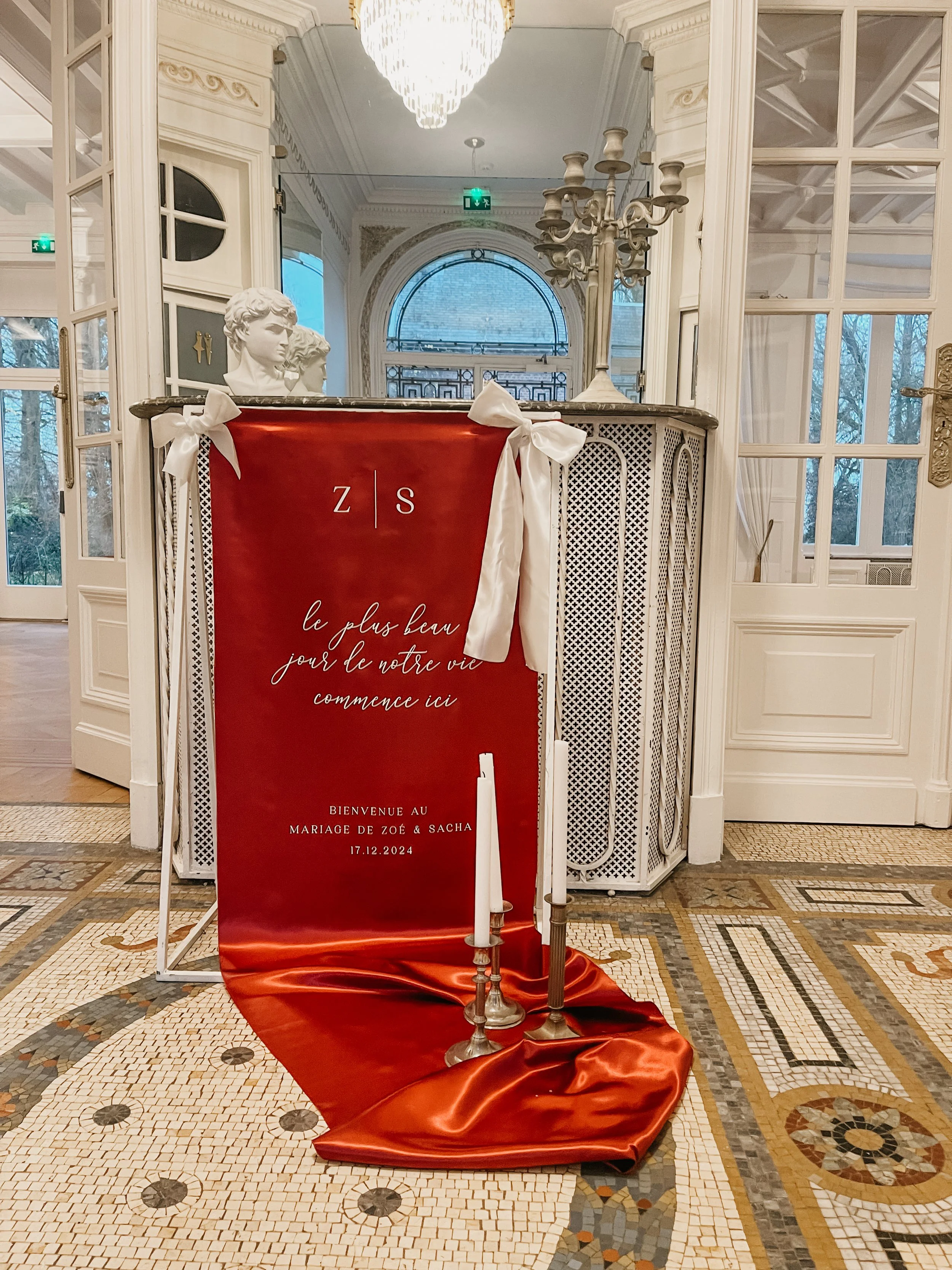 Panneau rouge avec une inscription en français pour un mariage, décoré de rubans blancs et de bougies, situé dans un intérieur élégant avec chandelier en cristal et statues en marbre.