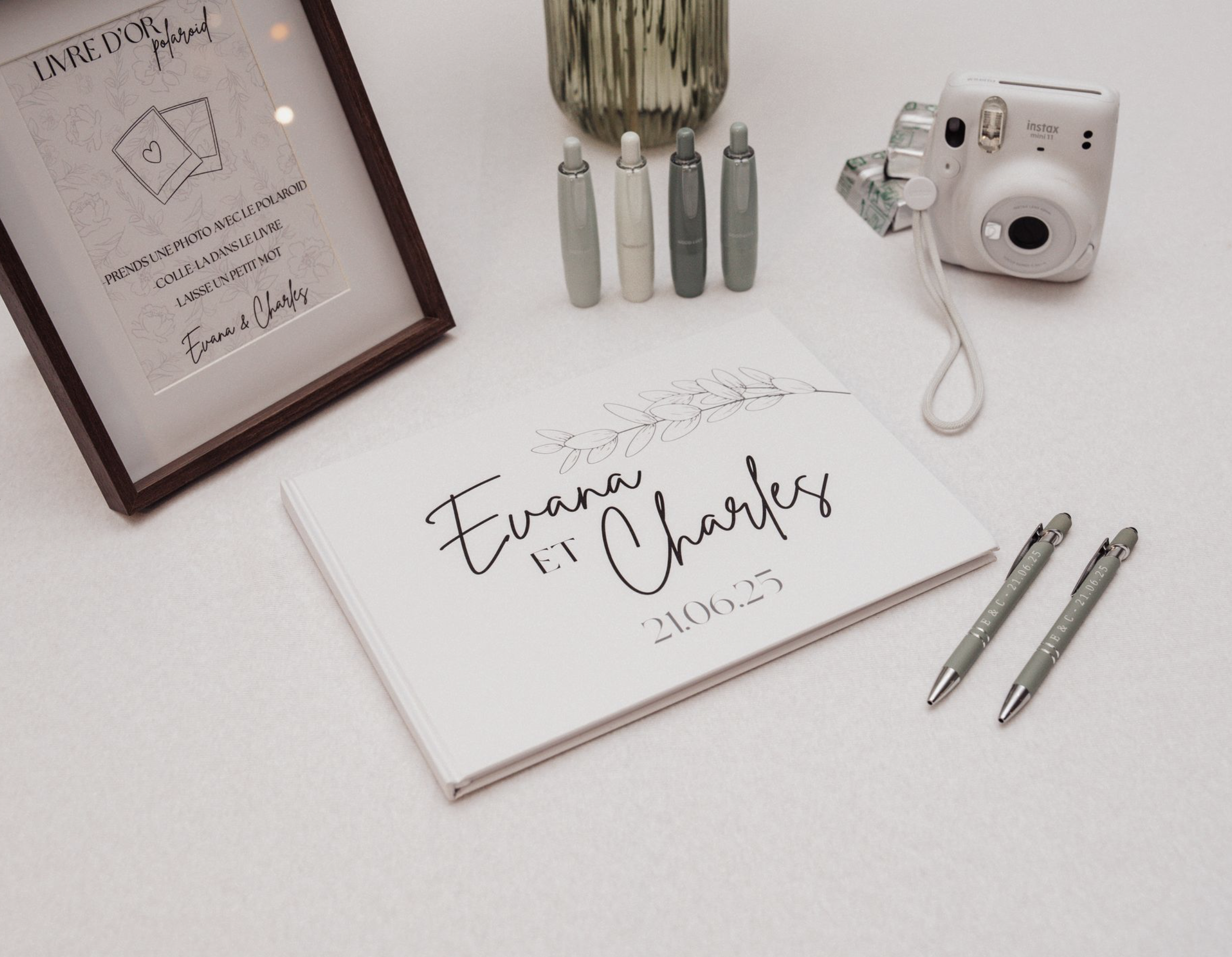 Mise en place de préparatifs pour un mariage avec un livre d'or, un tableau, des stylos, un appareil photo Polaroid, et des petits flacons, sur une table blanche.