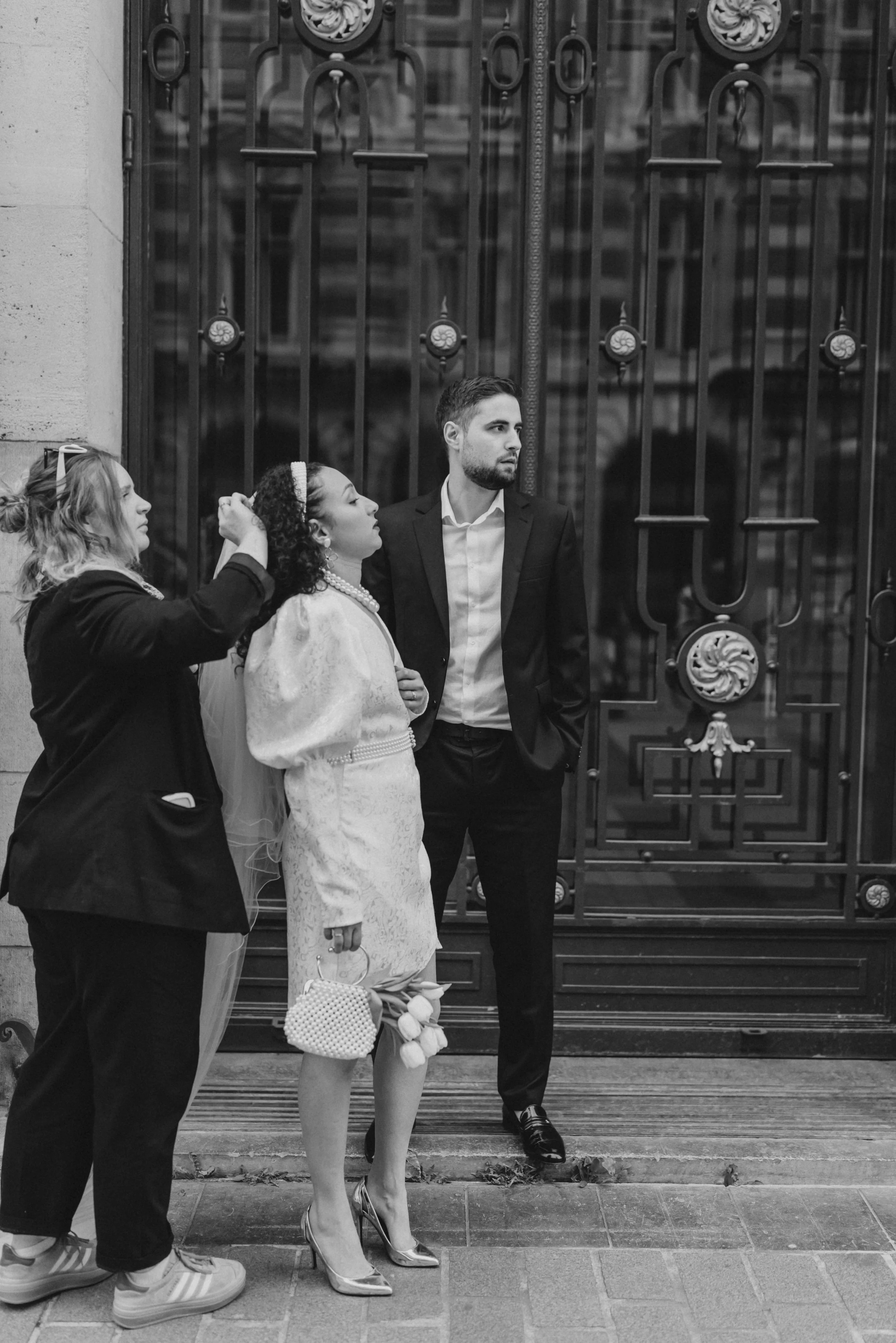 Une femme en robe élégante, tenant un bouquet de fleurs, se fait coiffer par une femme à gauche, tandis qu'un homme en costume noir se tient à côté devant une grille en fer ornée d'éléments décoratifs, dans une scène d'extérieur en noir et blanc.