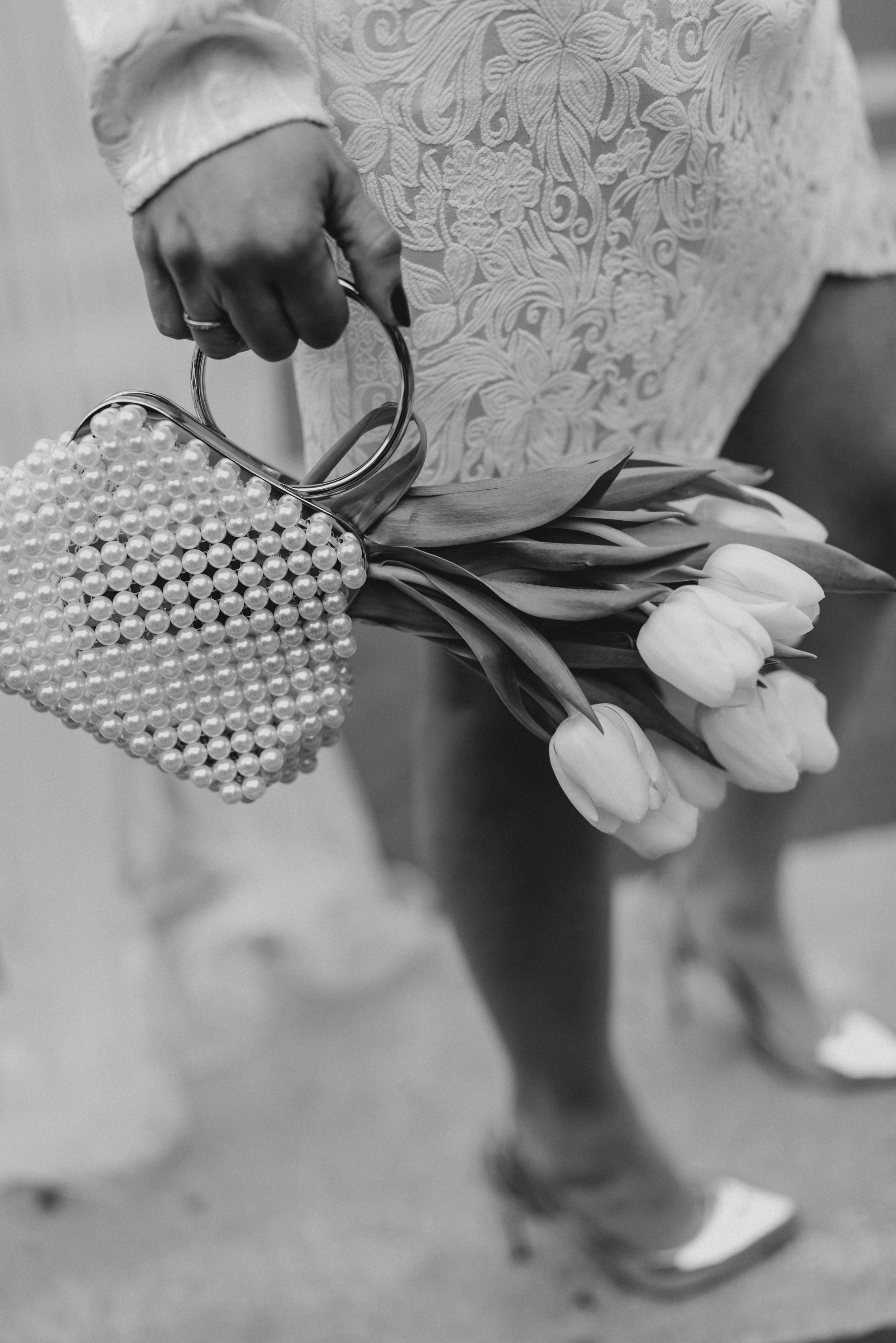 Une main portant une bague tient un sac en forme de cœur orné de perles, contenant des tulipes blanches, avec une personne portant une robe en dentelle et des chaussures à talons en arrière-plan.