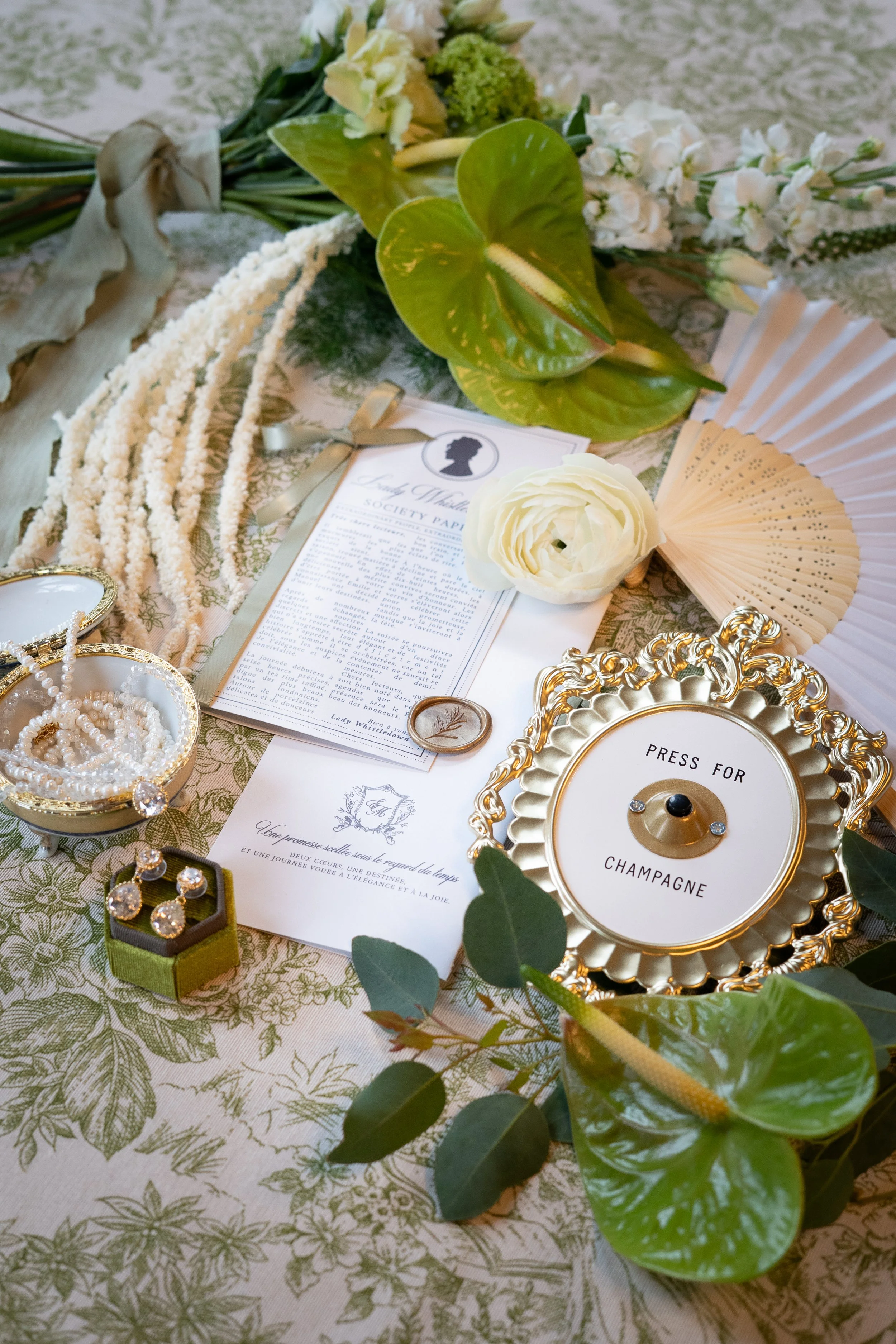 Napperon floral avec décoration de bijoux, fleurs blanches et accessoires pour une table de fête ou mariage, dont un panneau 'Press for Champagne', papiers et fleurs vertes.