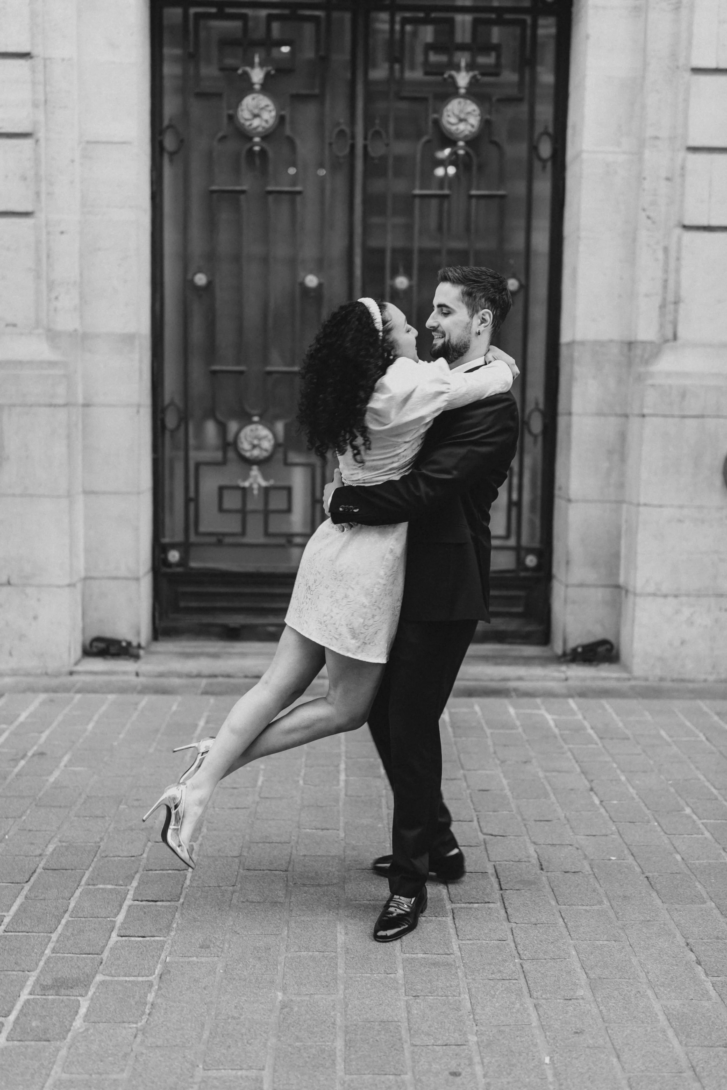 Un couple danse devant une grande porte en arrière-plan, l'homme en costume sombre et la femme en robe courte claire, ils se regardent et s'embrassent tendrement.