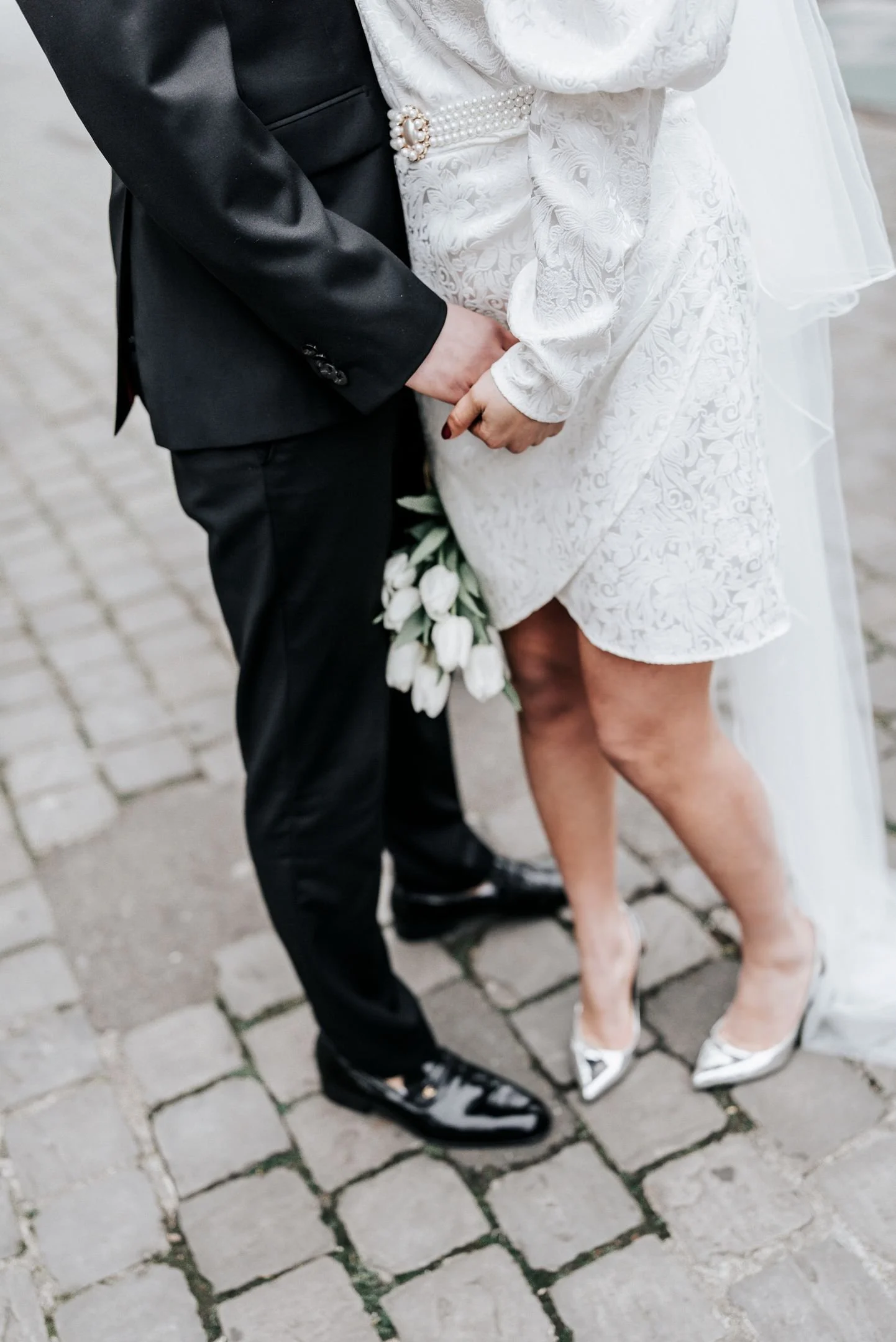 Un couple de mariage, tenant la main, le marié en costume noir et la mariée en robe blanche avec des chaussures à talons argentées, tenant un bouquet de tulipes blanches, sur un pavé en extérieur.