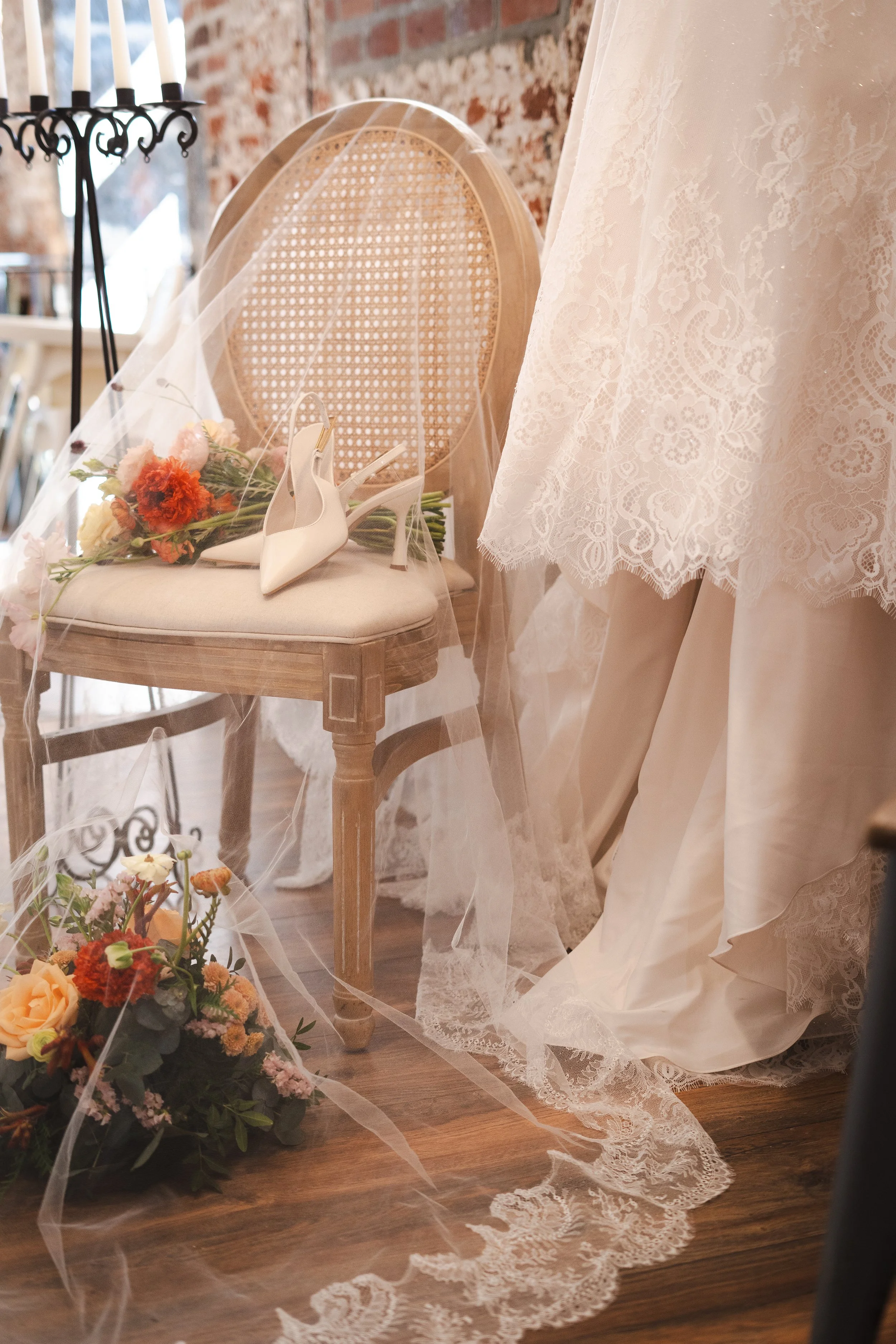 Une chaise en bois avec un coussin et un voile de mariée qui y est posé, entourée de bouquets de fleurs colorées, une robe de mariée en dentelle blanche est suspendue à droite et un chandelier à bougies en arrière-plan.