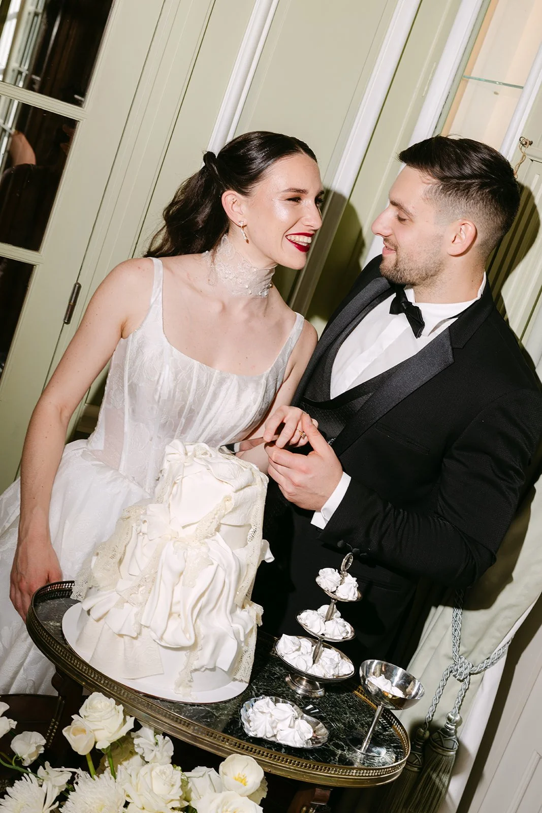 Un couple de mariés partage un moment heureux lors de leur mariage, avec un gâteau blanc décoré, dans une pièce élégante.