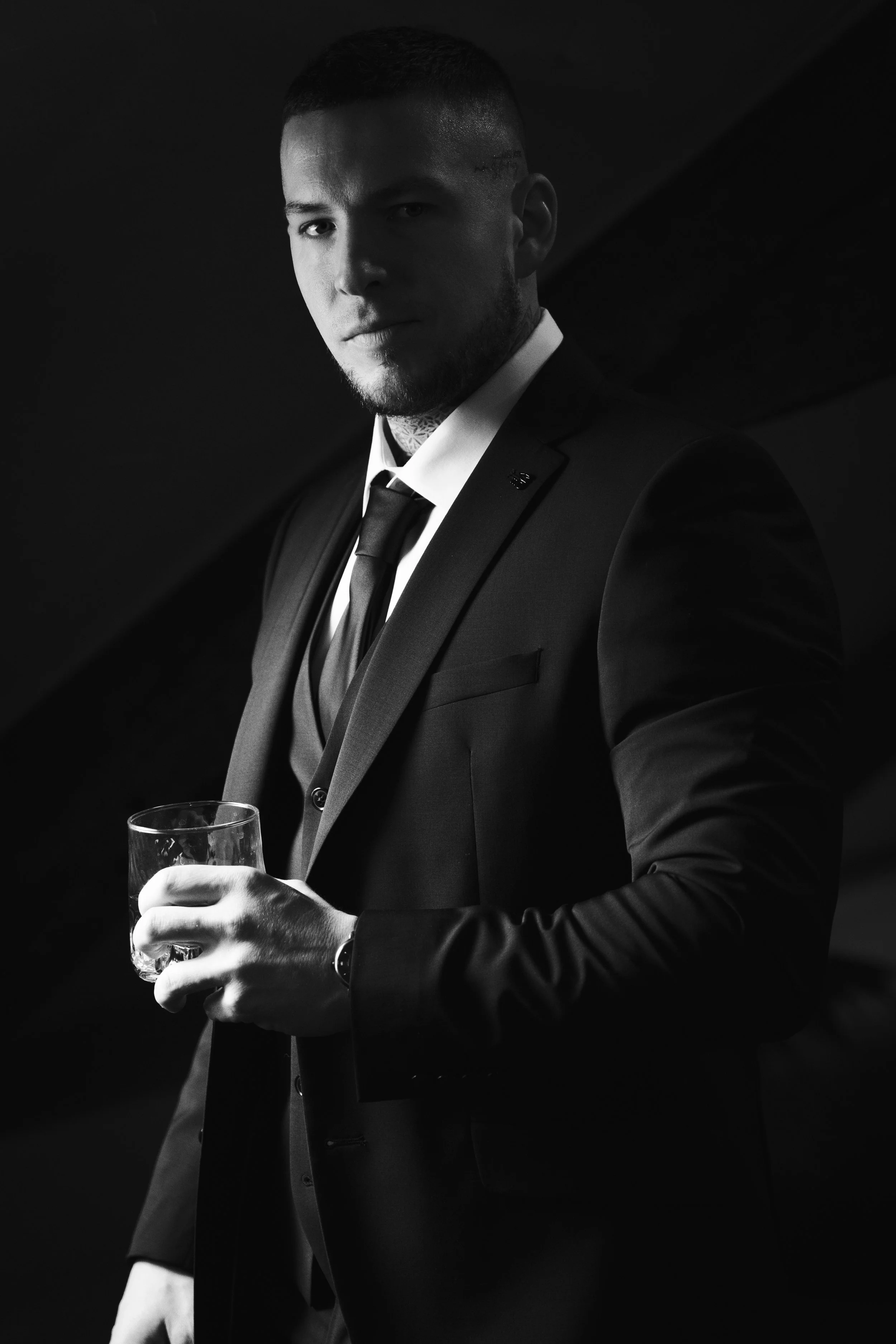 Un homme habillé en costume noir, tenant un verre à whisky, dans une ambiance sombre et élégante.