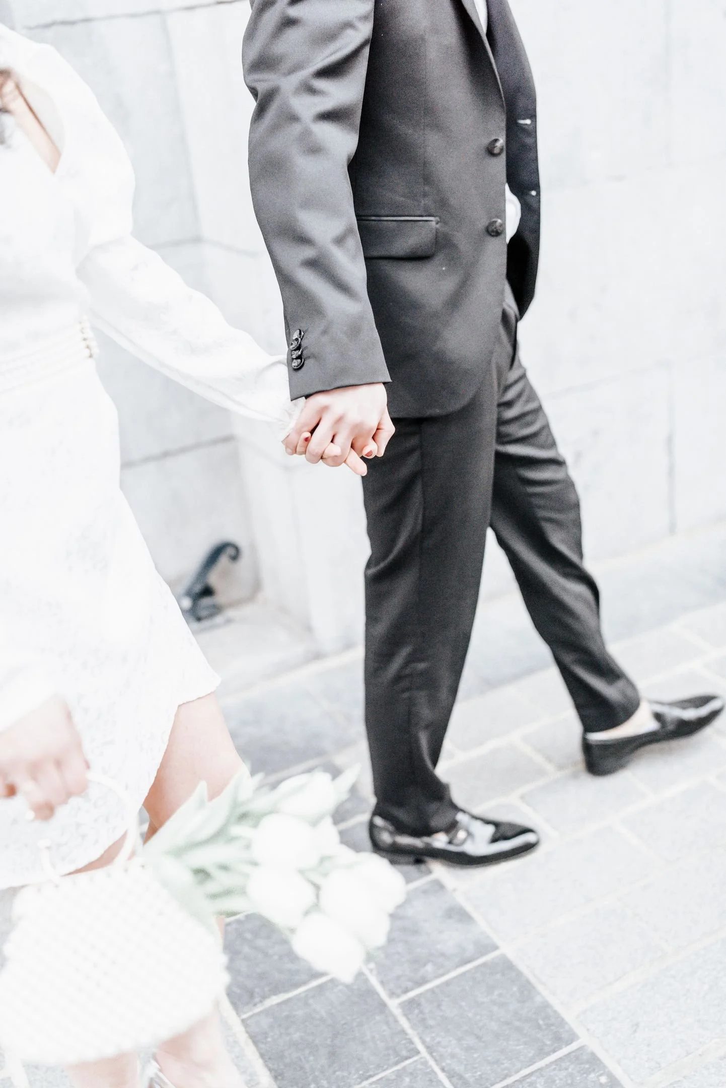 Un couple tenant la main lors d'une cérémonie, portant des vêtements formels, devant un mur en pierre.