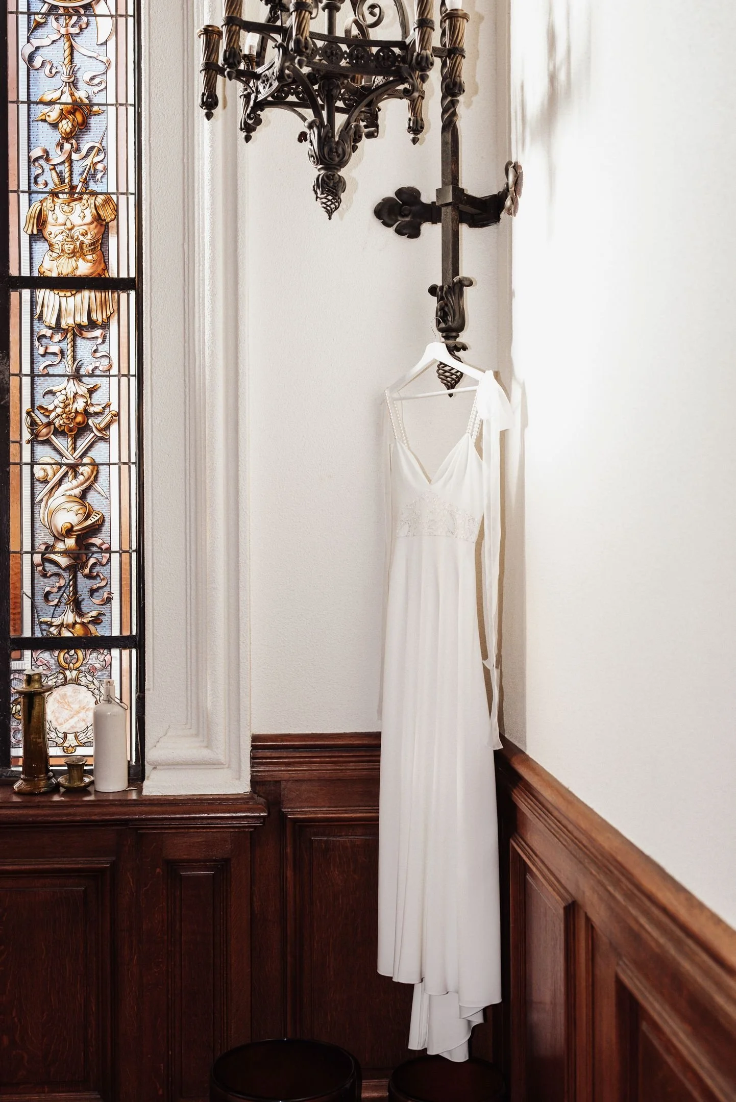 Robe de mariée blanche suspendue à un crochet sur un mur dans une pièce élégante avec boiseries en bois foncé et vitrail coloré.