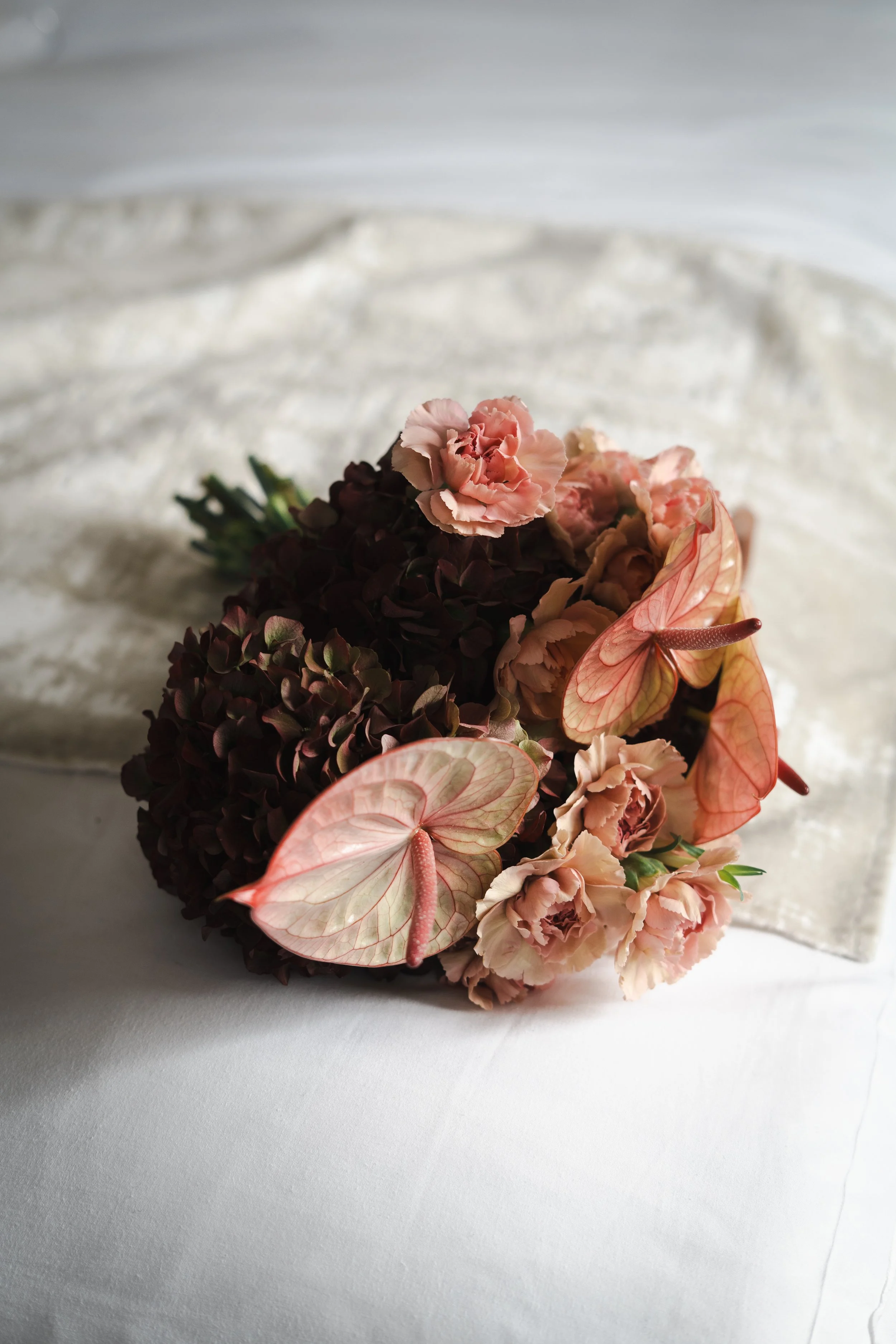 Un bouquet de fleurs composé de pivoines roses, de hortensias sombres et de feuilles de caladium rose avec des veines roses, posé sur une surface blanche.