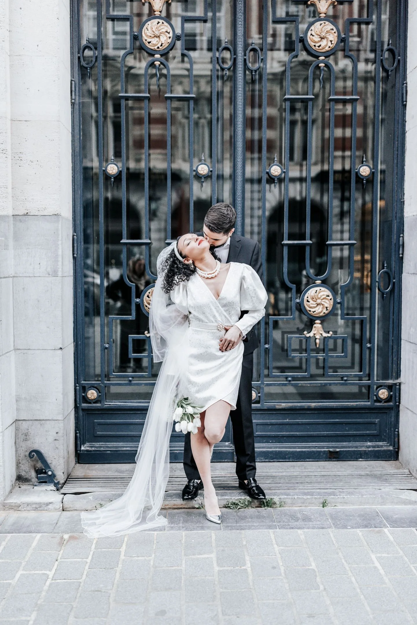 Un couple lors d'un mariage, la femme en robe blanche et le mari en costume noir, posant devant une grande porte en fer ornée de détails dorés.