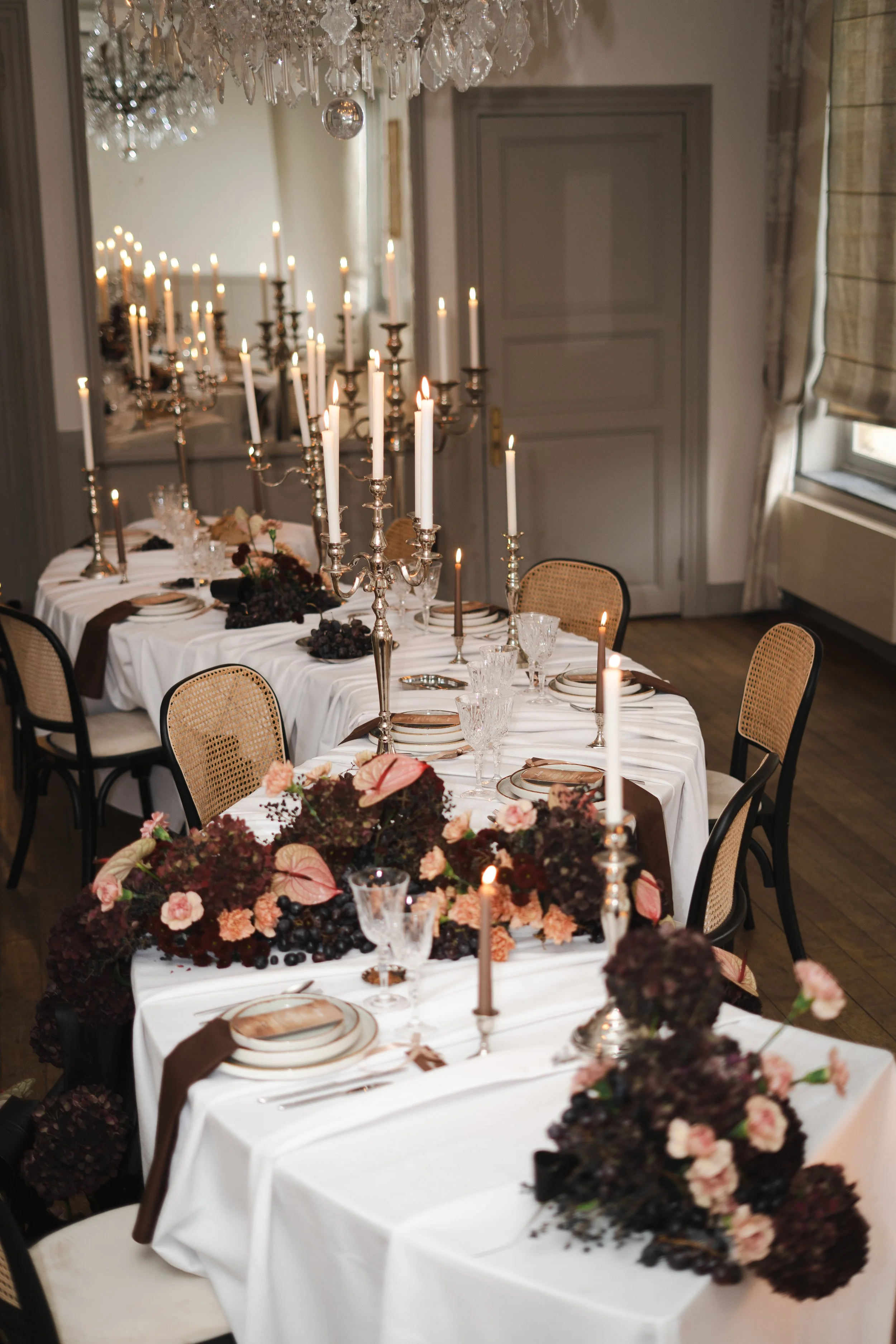 Une salle à manger élégante avec une grande table couverte d'une nappe blanche, décorée avec des chandeliers en argent, des fleurs foncées et des fruits, pour un repas sophistiqué.