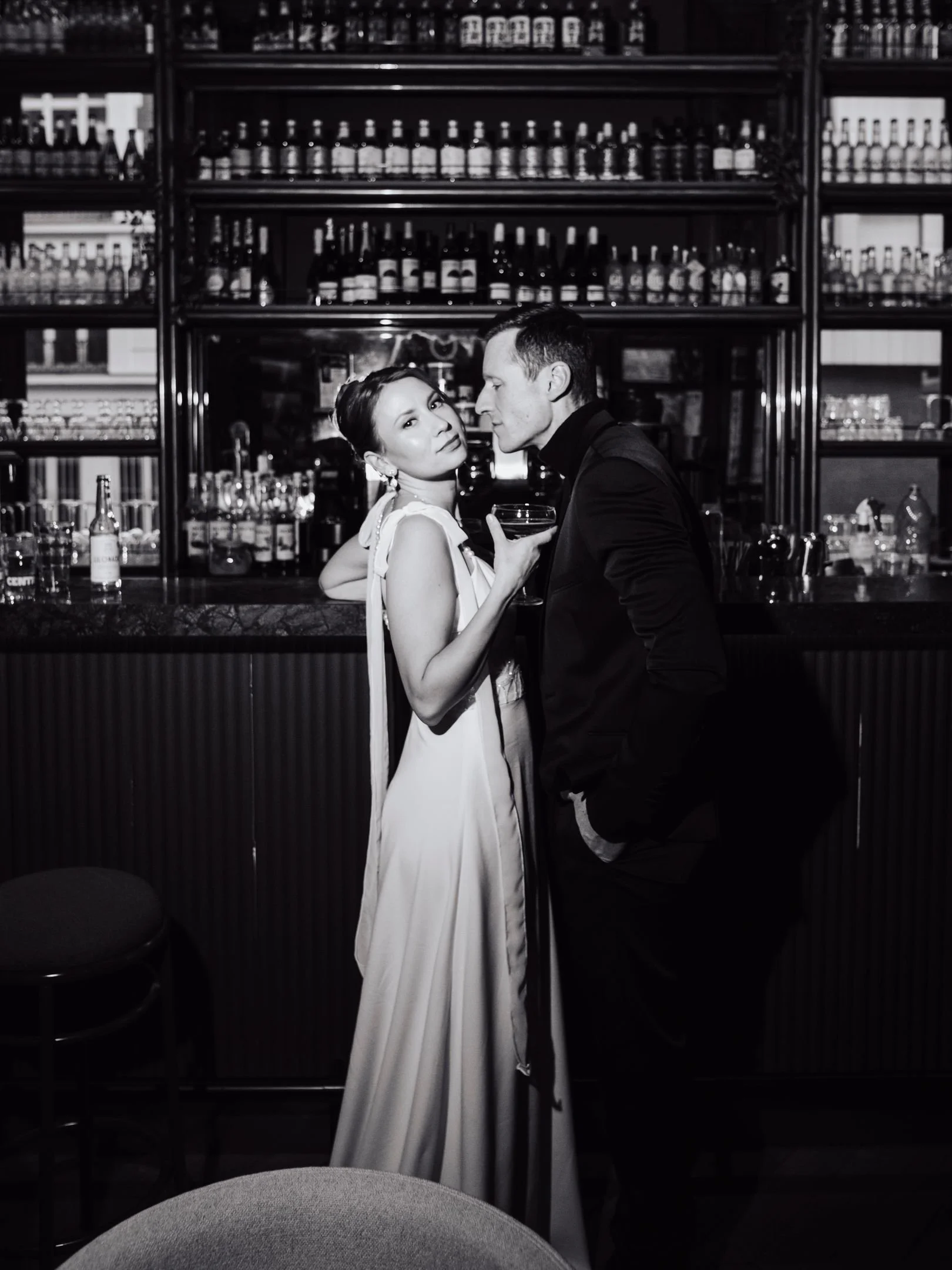 Un homme et une femme élégamment habillés posent au bar, la femme tient un verre à cocktail et regarde vers l'objectif, le bar en arrière-plan est bien approvisionné en bouteilles.