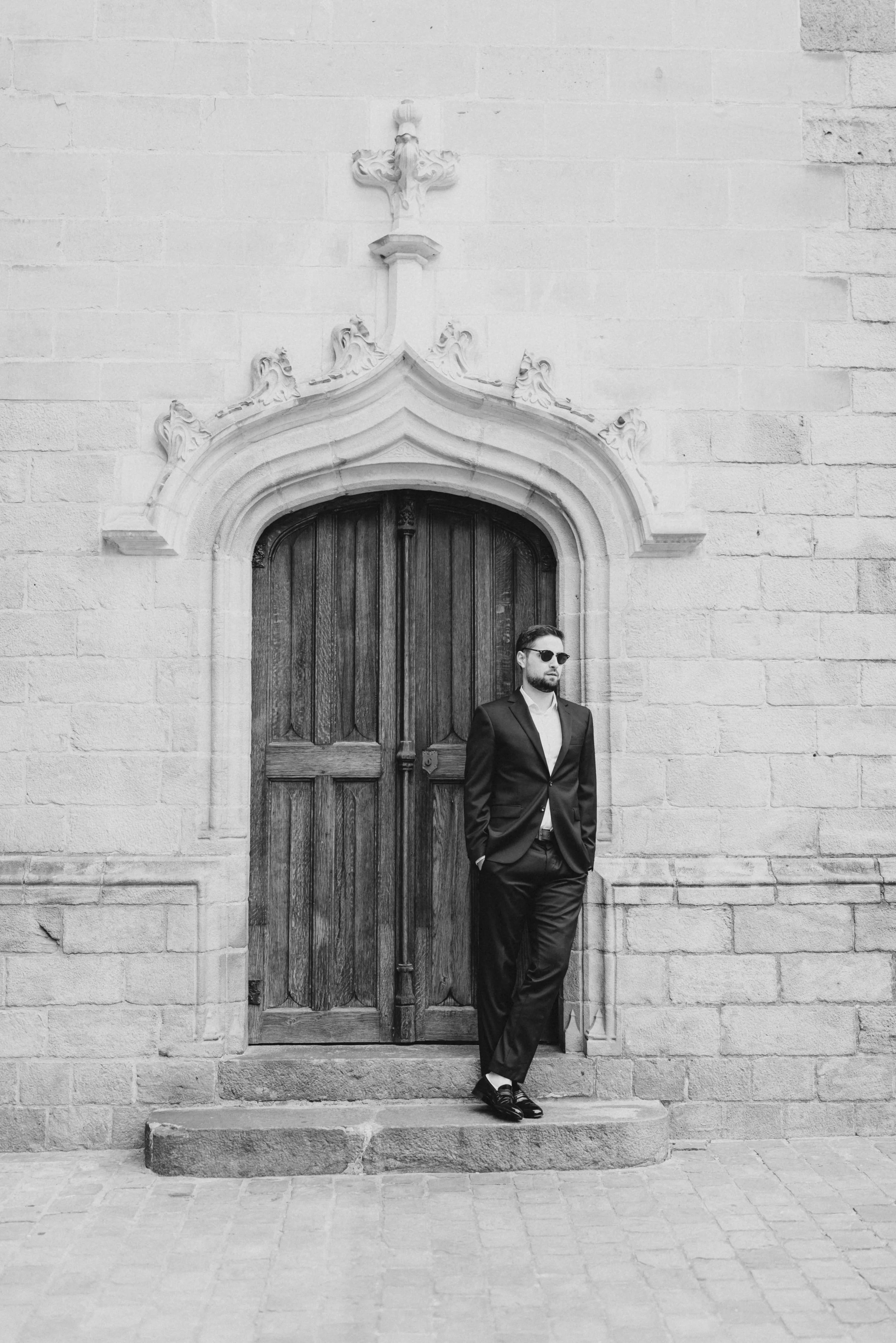 Homme en costume sombre portant des lunettes de soleil, debout devant une porte en bois ancienne encadrée d'une architecture gothique sur un mur en pierre, photographie en noir et blanc.