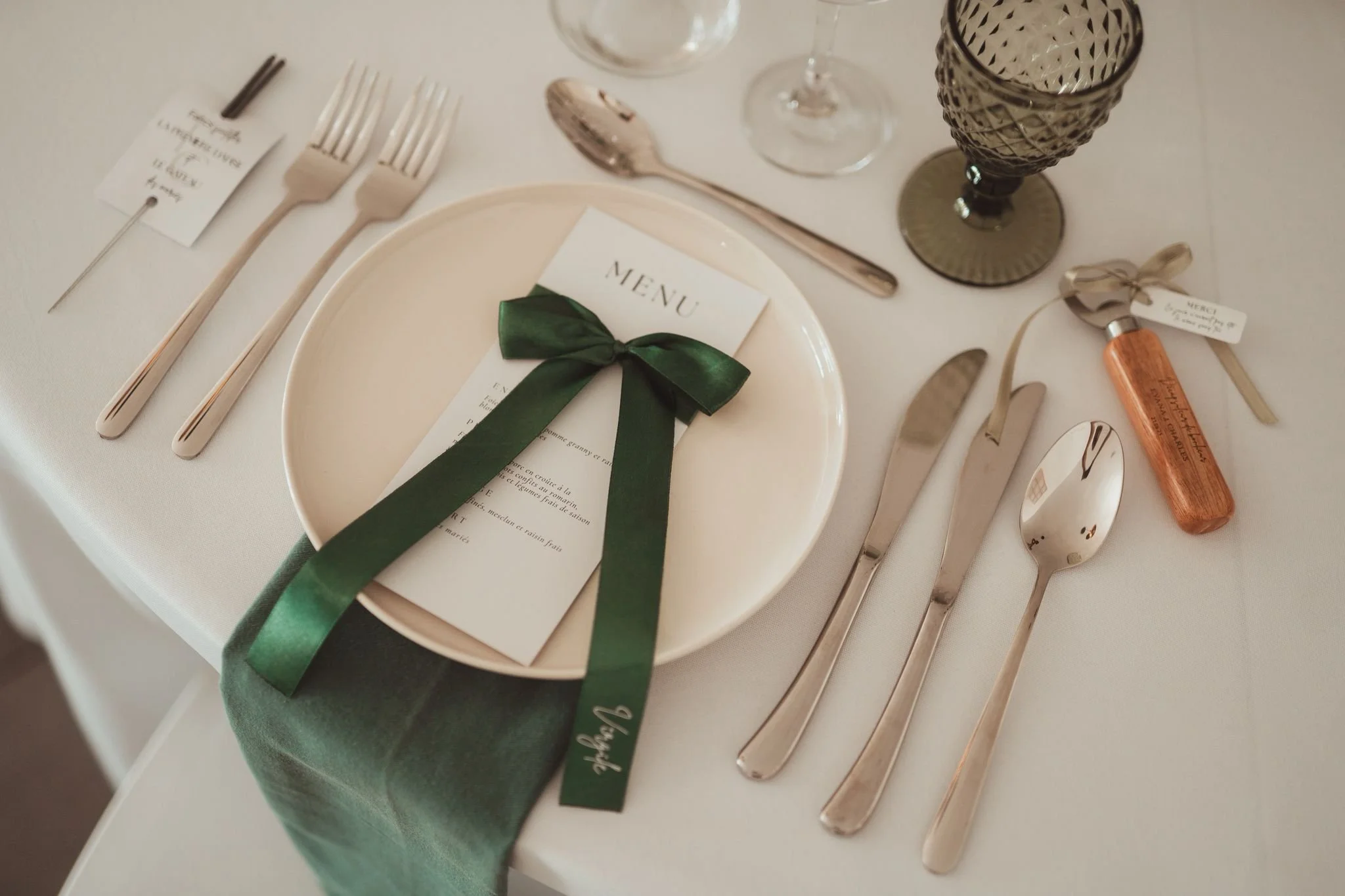 Plats de table élégants avec assiette, menu avec ruban vert, couverts en argent, verres à vin, pièce de service en bois, et marque-place, décor de mariage sophistiqué.