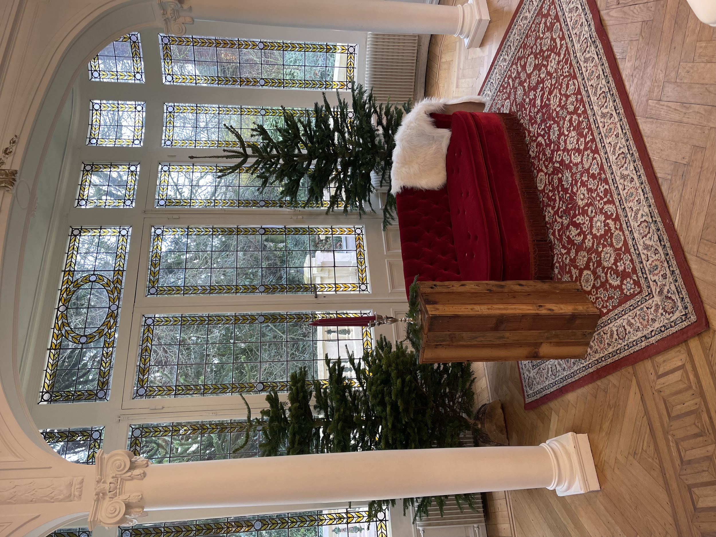 Un salon décoré pour Noël avec un sapin d'environ 3 mètres, un canapé en velours rouge, un tapis oriental, et une grande fenêtre avec des vitraux.