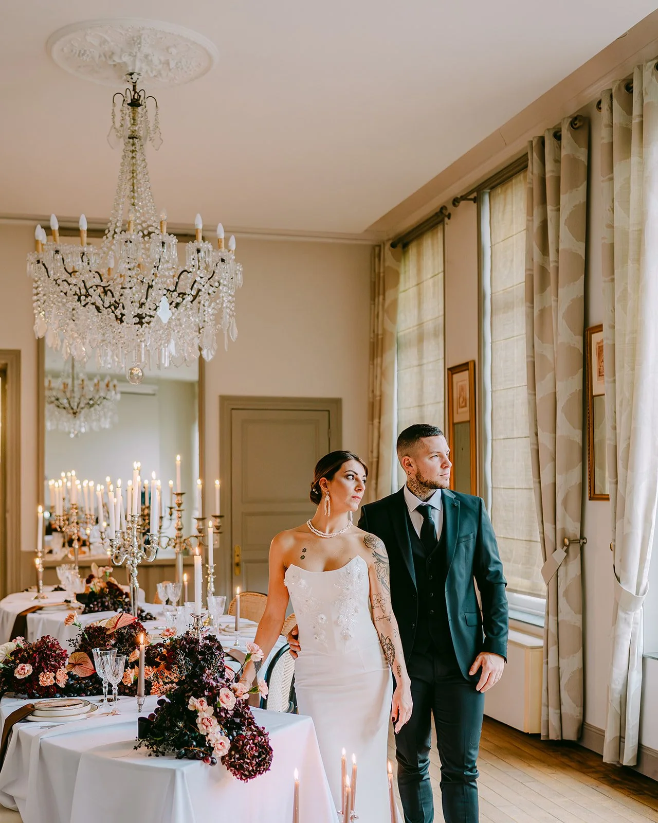 Un couple habillé pour un mariage dans une salle décorée avec des chandeliers, des fleurs sombres et des bougies.
