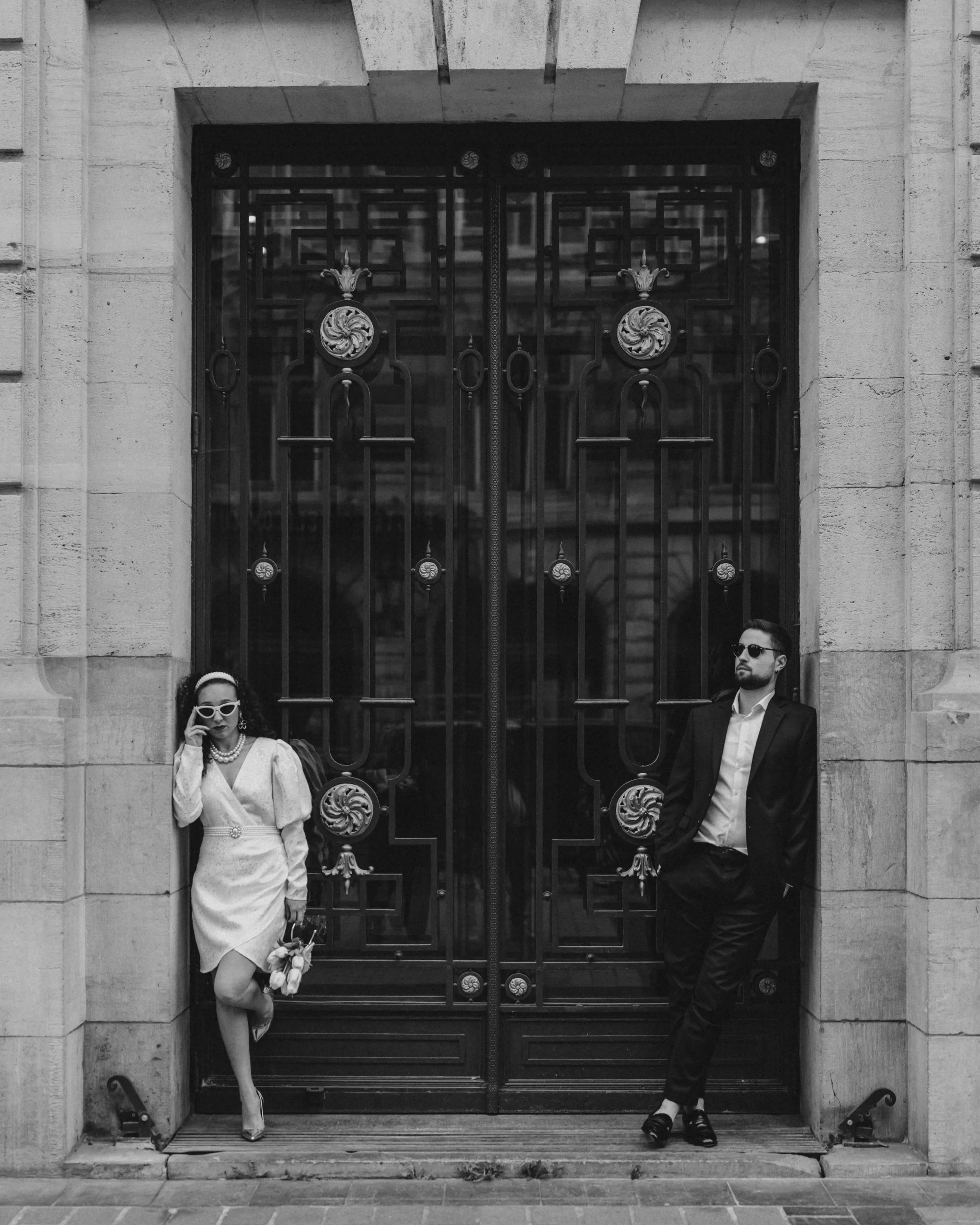Deux personnes habillées élégamment, une femme à gauche portant une robe blanche, des lunettes de soleil, et tenant un bouquet, un homme à droite en costume noir, tous deux posé devant une porte en métal ornée d'un bâtiment en pierre.