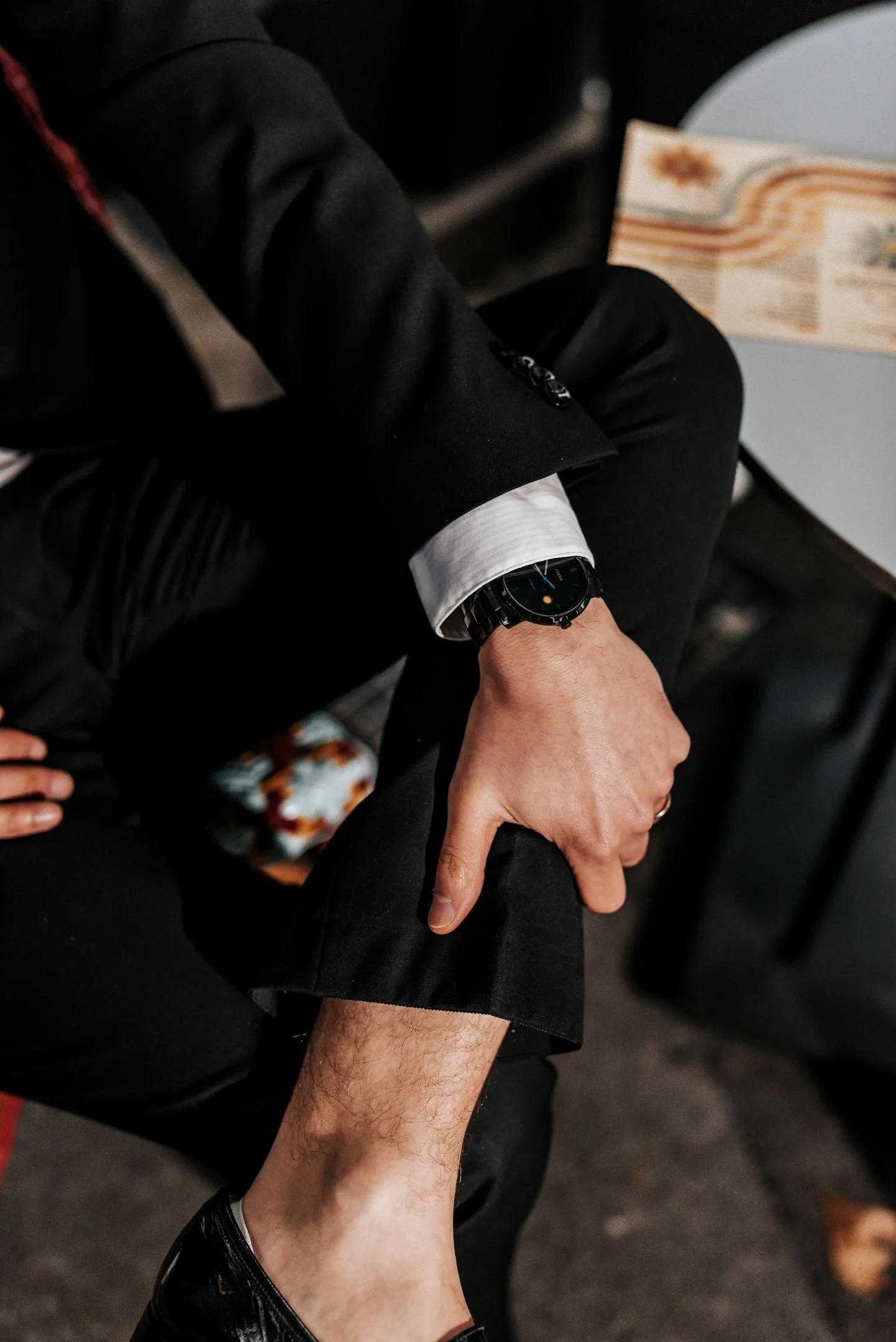 Une personne habillée d'un costume noir regarde sa montre, assise à une table avec un gâteau.