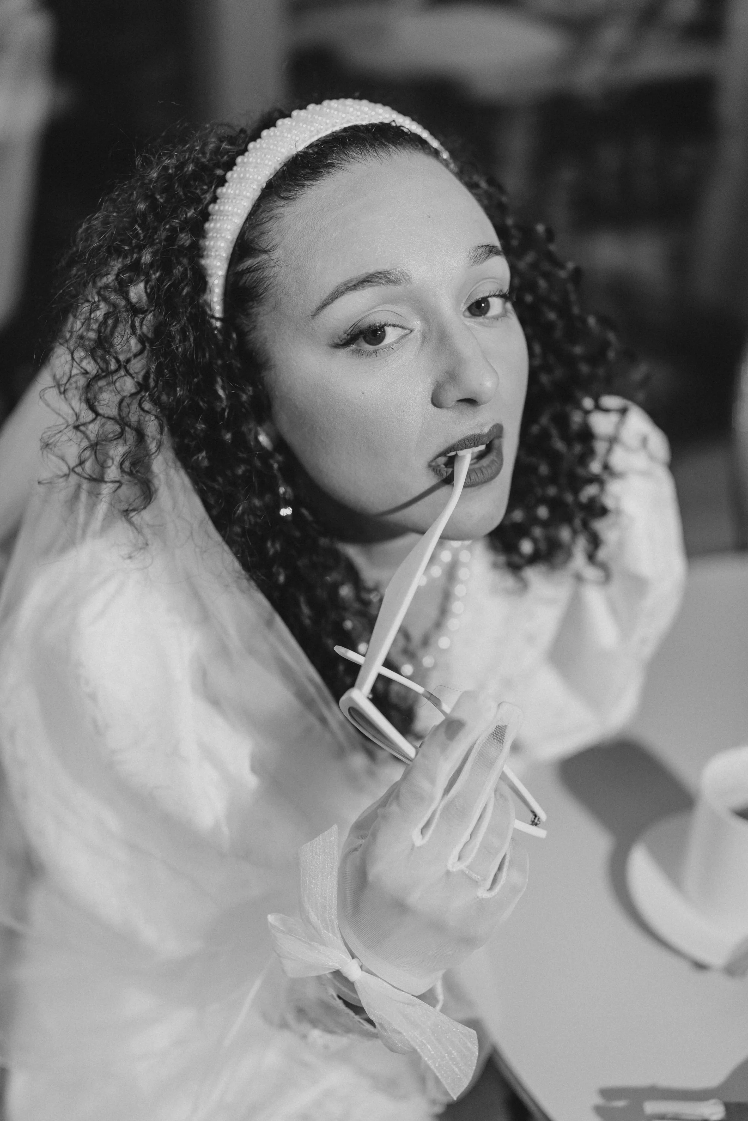 Une femme aux cheveux bouclés porte un bandeau de perles, tient des lunettes, et mord une paille sur le rebord de sa cuillère, regardant vers l'objectif.