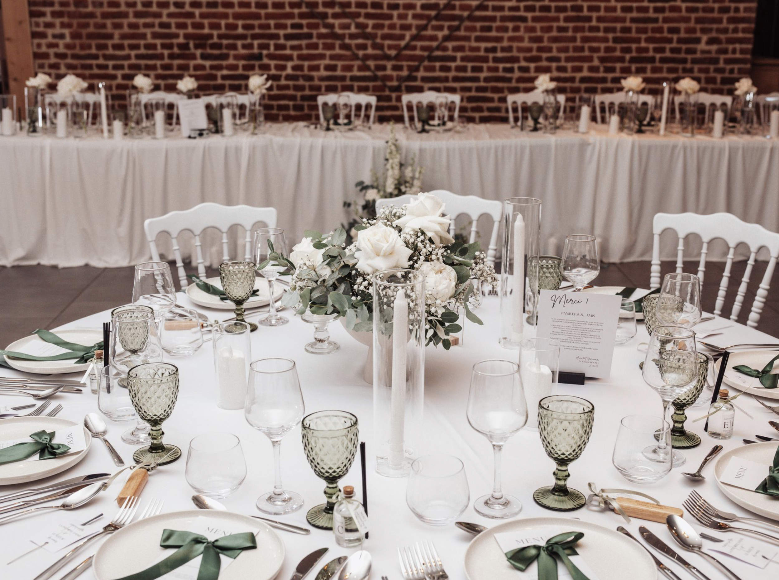 Table de mariage élégante avec une grande composition florale blanche centrale, bougies, verres à vin vert, assiettes avec des nœuds verts, et menu. En arrière-plan, longue table décorée, avec chaises blanches et bougies, contre un mur en briques.