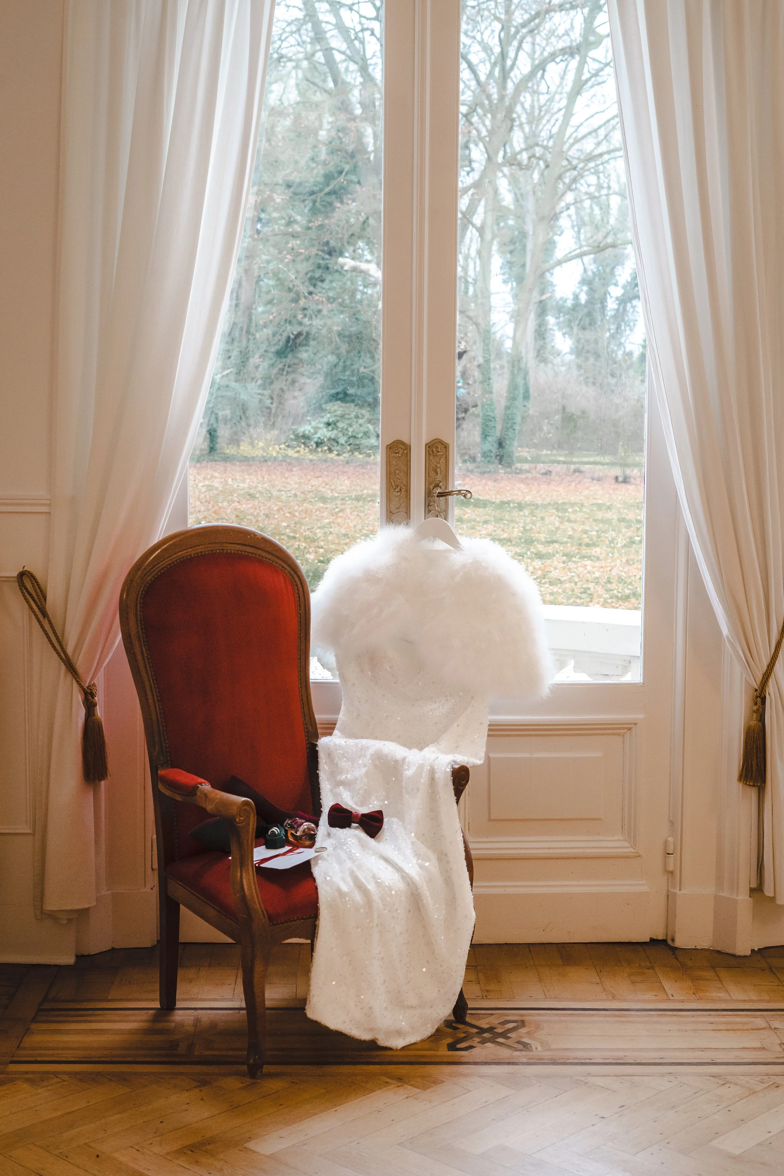 Une robe de mariée blanche en chiffon avec un voile en plumes est suspendue sur un cintre derrière une fenêtre, avec une chaise en bois rouge face à la fenêtre, dans une pièce élégante avec des rideaux blancs. Il y a des petits accessoires posés sur 