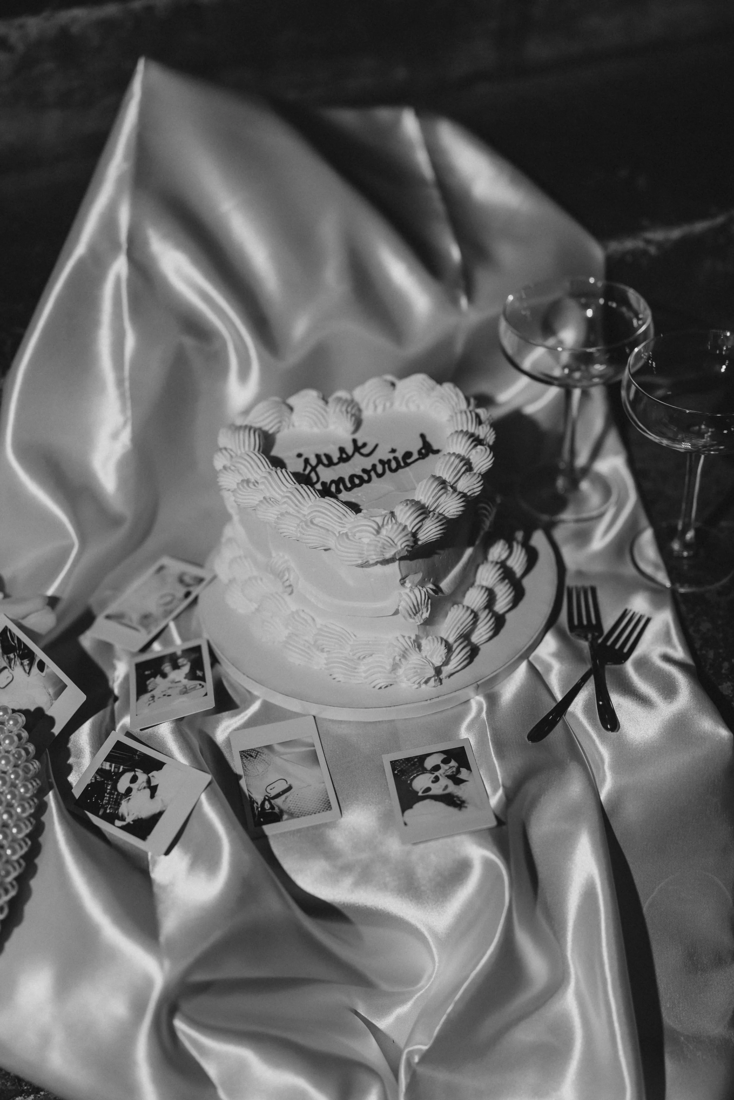 Gâteau de mariage avec le message 'just married' écrit dessus, entouré de chrétiens à glaçage, sur une table en satin avec des flûtes à champagne, des fourchettes, des photos de mariage.