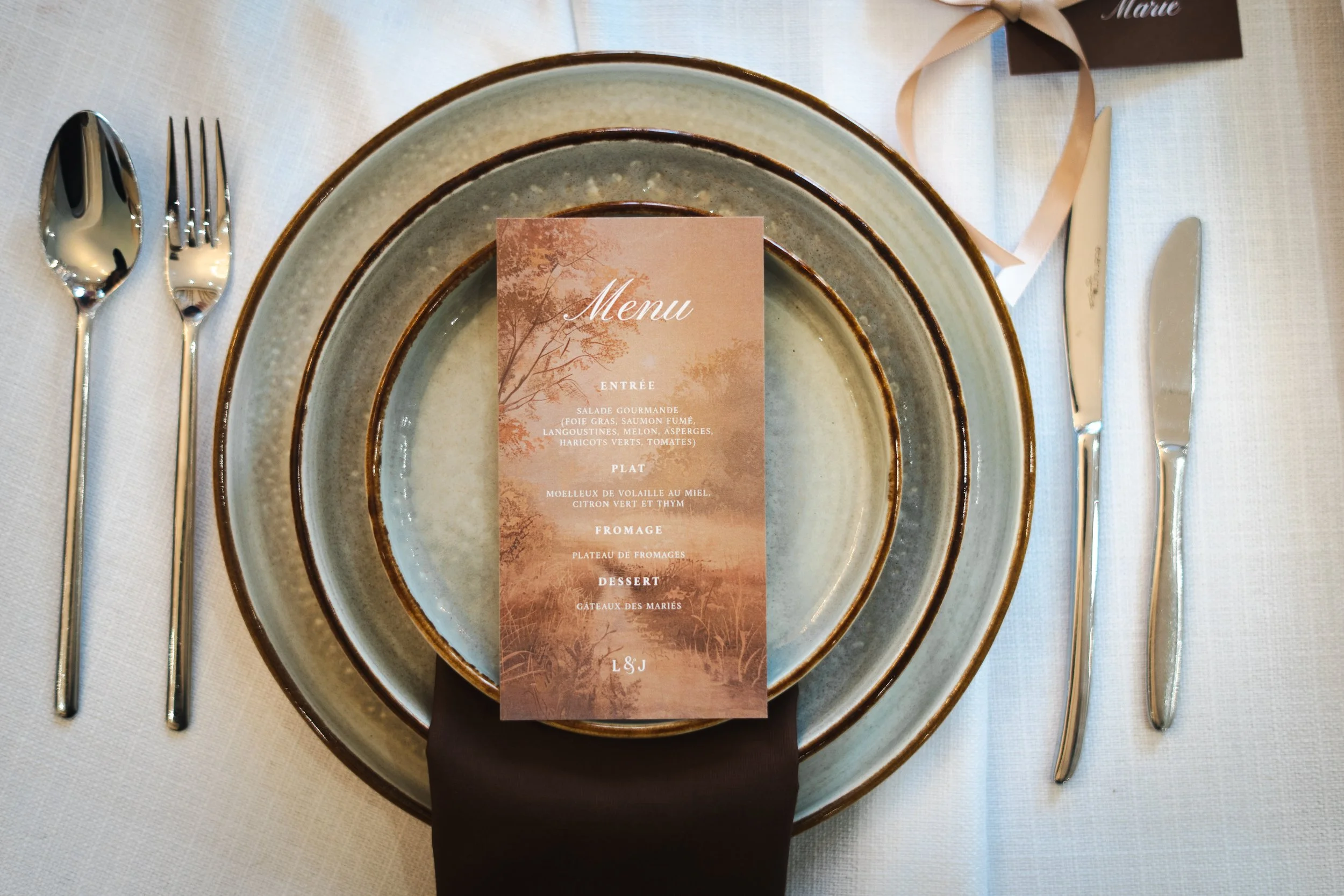 Une table de dîner élégante avec deux assiettes empilées, un menu posé dessus, une fourchette, un couteau, une cuillère en métal argenté, et une nappe blanche. Le menu est rose avec un paysage dessiné et comporte plusieurs catégories de plats.