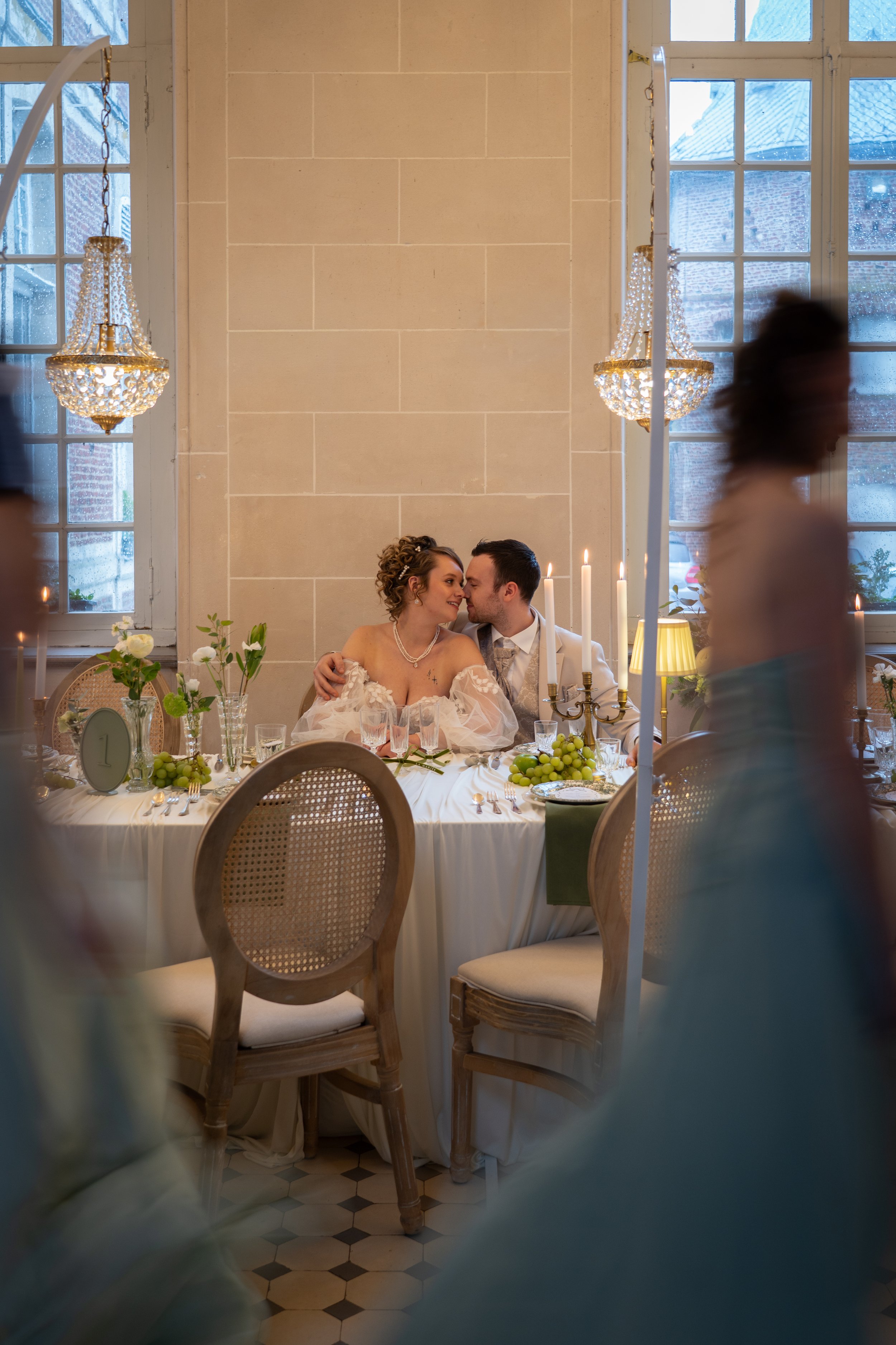 Un couple de mariés s'embrassant à une table de mariage élégante avec bougies, fleurs, et raisins dans une salle éclairée par des lustres en cristal.