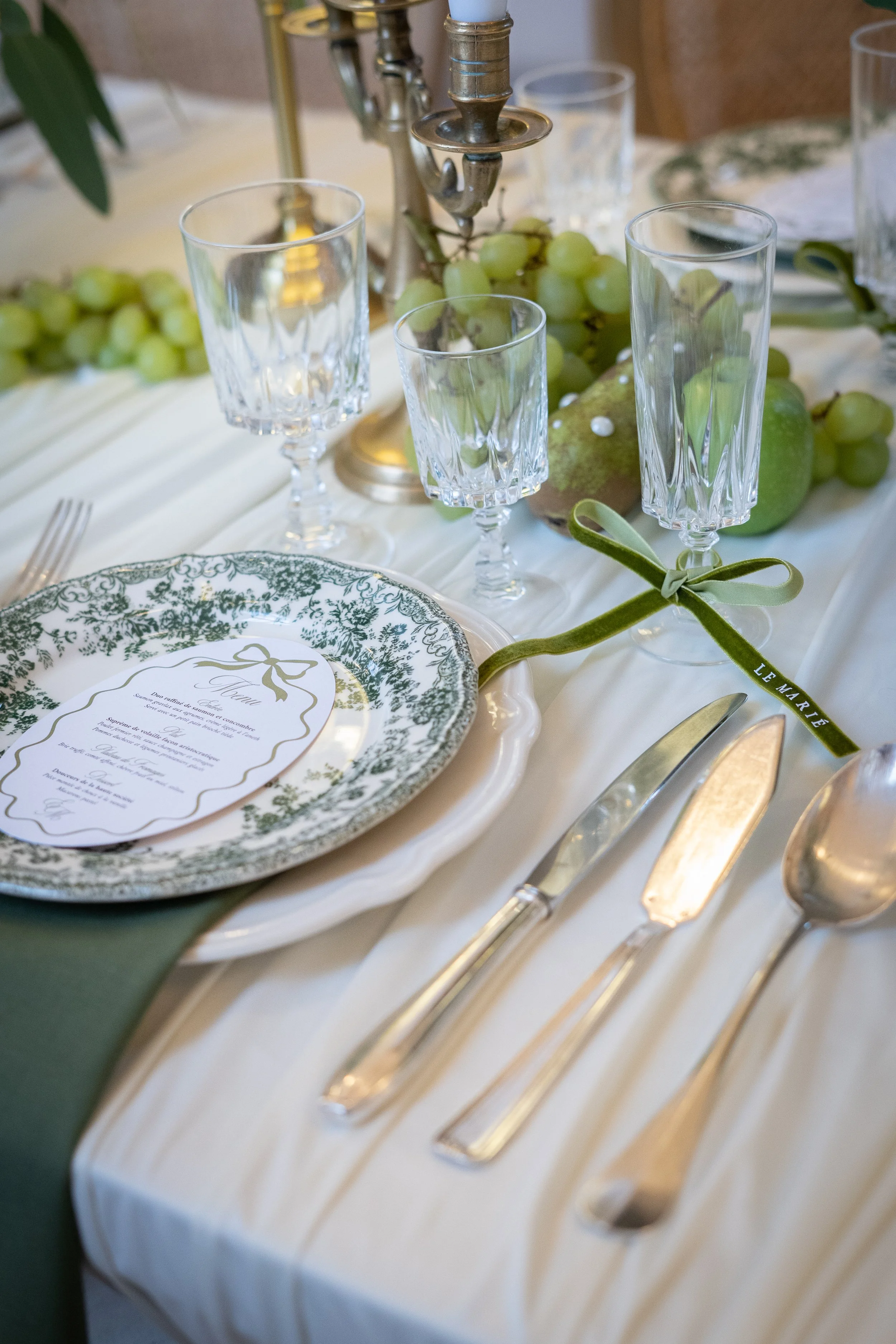 Une table de mariage élégante avec une assiette décorative, un menu, des couverts en argent, des verres en cristal, une guirlande de raisins verts, et un centre de table avec une bougie et une décoration en forme de branche avec un ruban vert portant