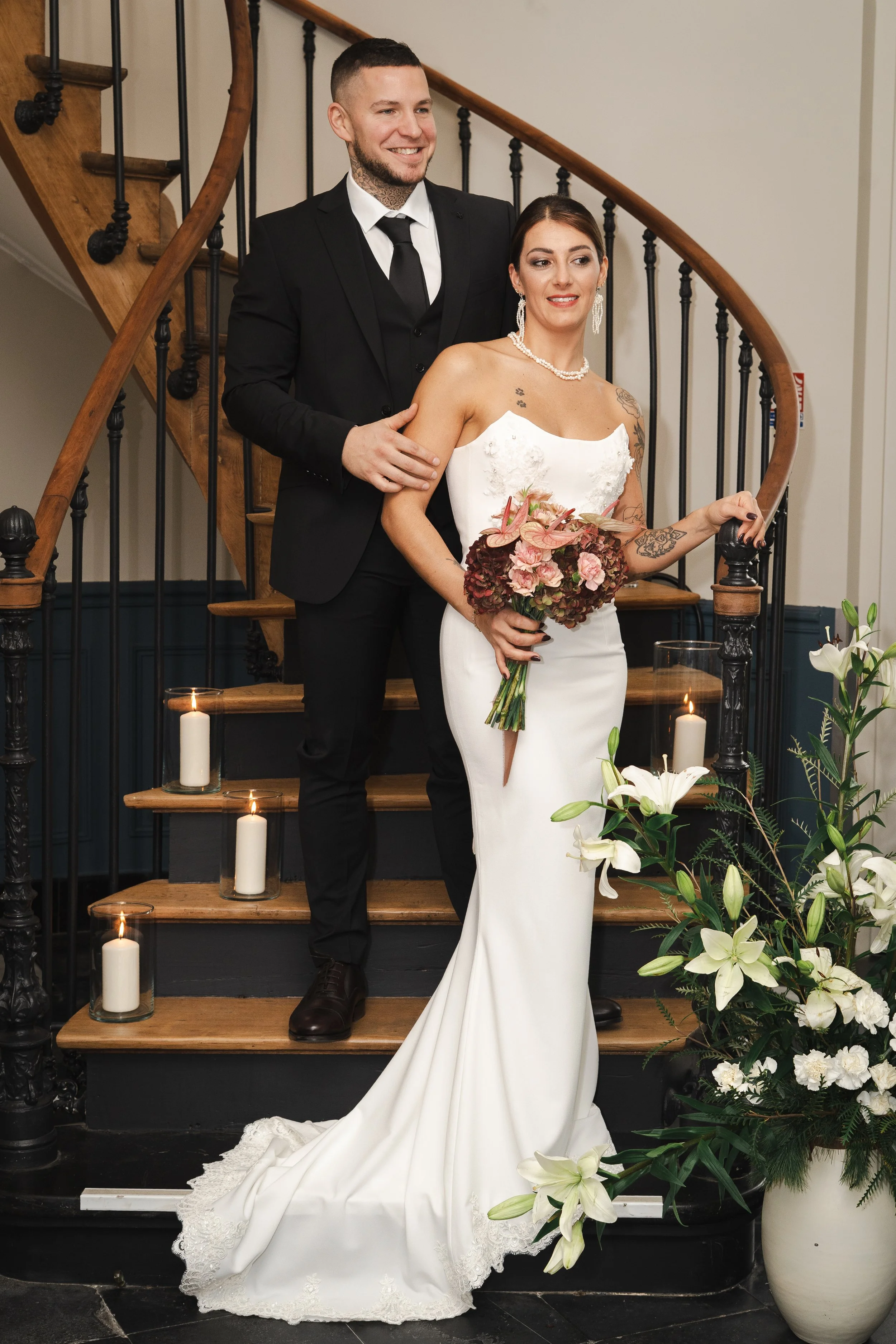 Un couple lors de leur mariage dans une maison, avec la mariée tenant un bouquet de fleurs roses et portant une robe de mariée blanche, le marié en costume noir, entourés de bougies et de fleurs blanches.