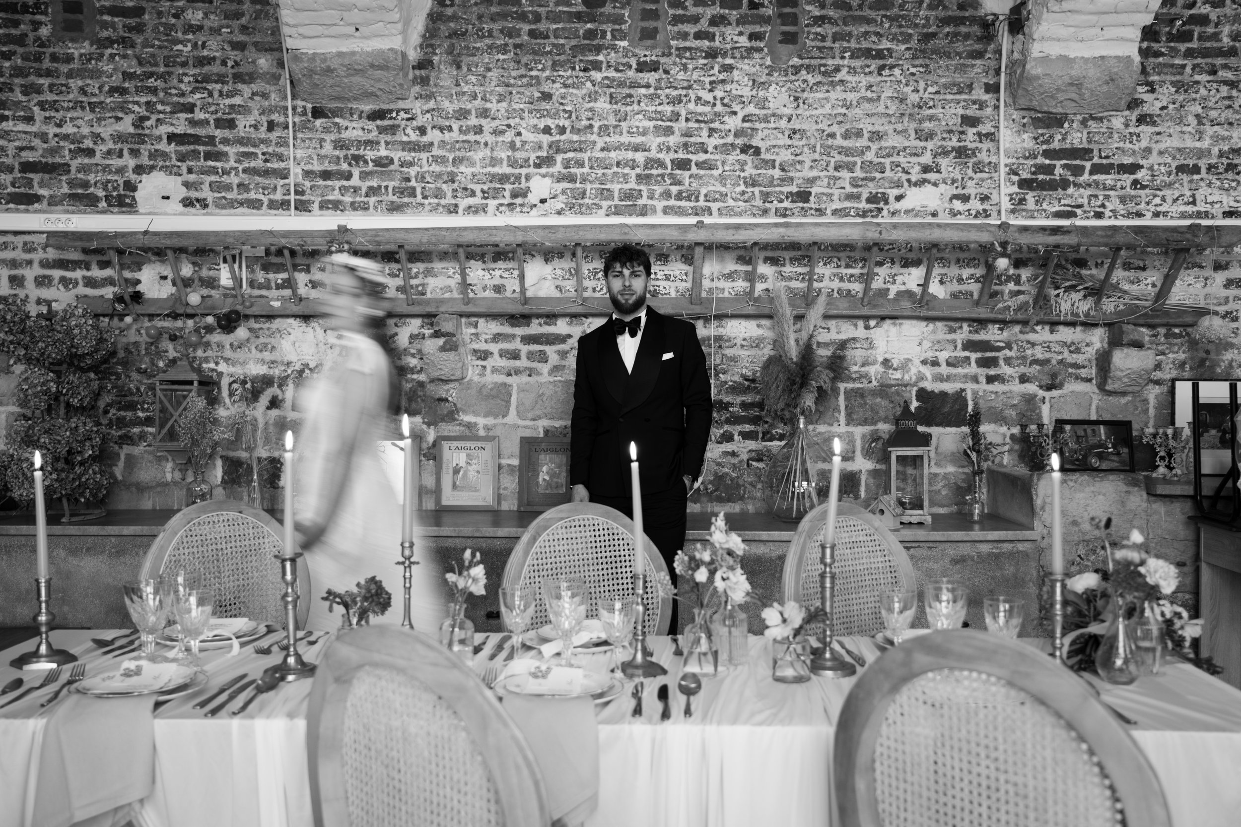 Un homme en costume noir et nœud papillon se tient derrière une table de repas décorée avec des bougies, des fleurs et des assiettes. La pièce a un mur en briques apparentes avec une étagère en bois au fond. Il y a aussi un verre de bouteille d'eau dans le décor.