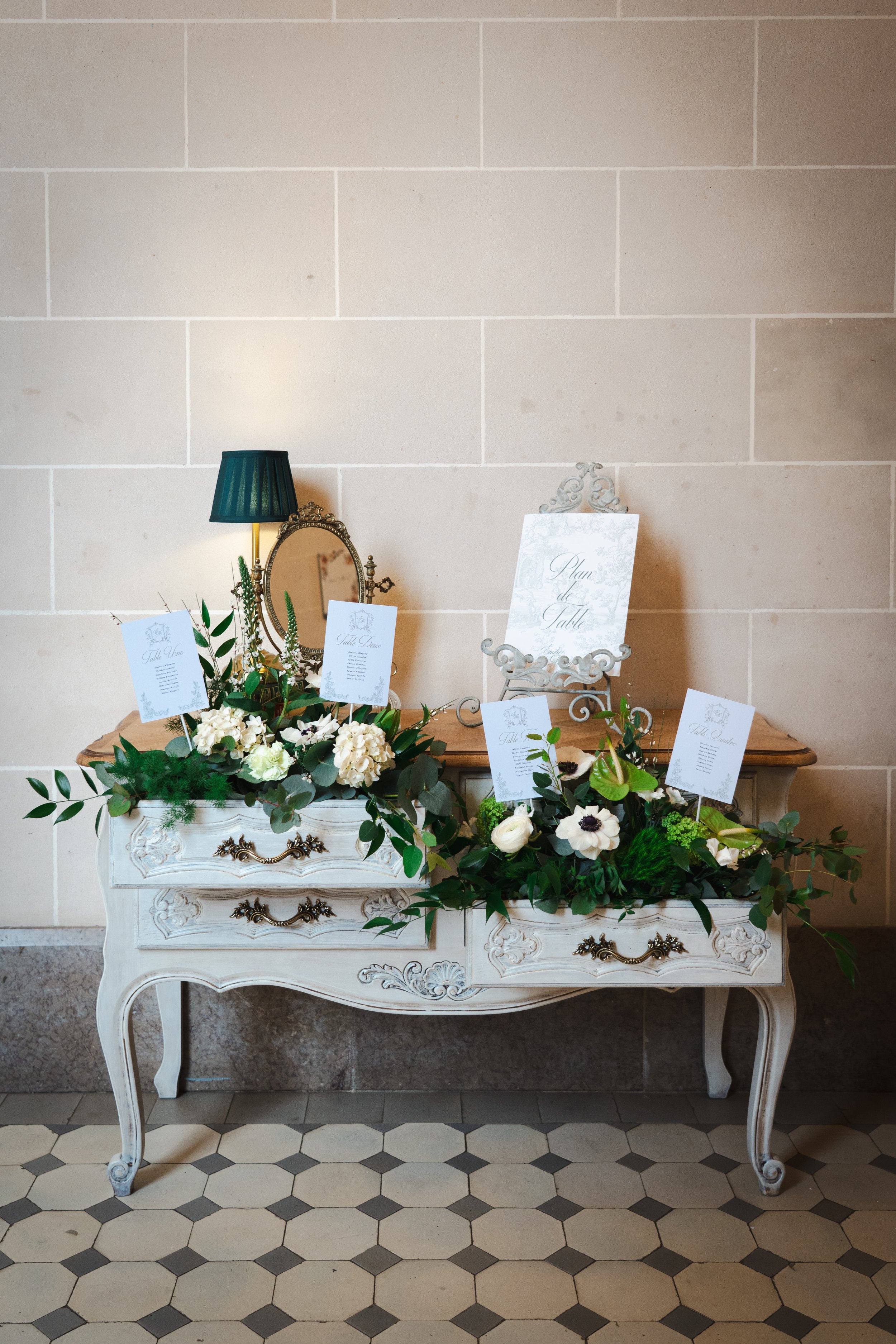 Une table de mariage décorée de fleurs blanches et vertes, avec des cartes de menu et une petite glace ancienne en fond, contre un mur en pierre beige.