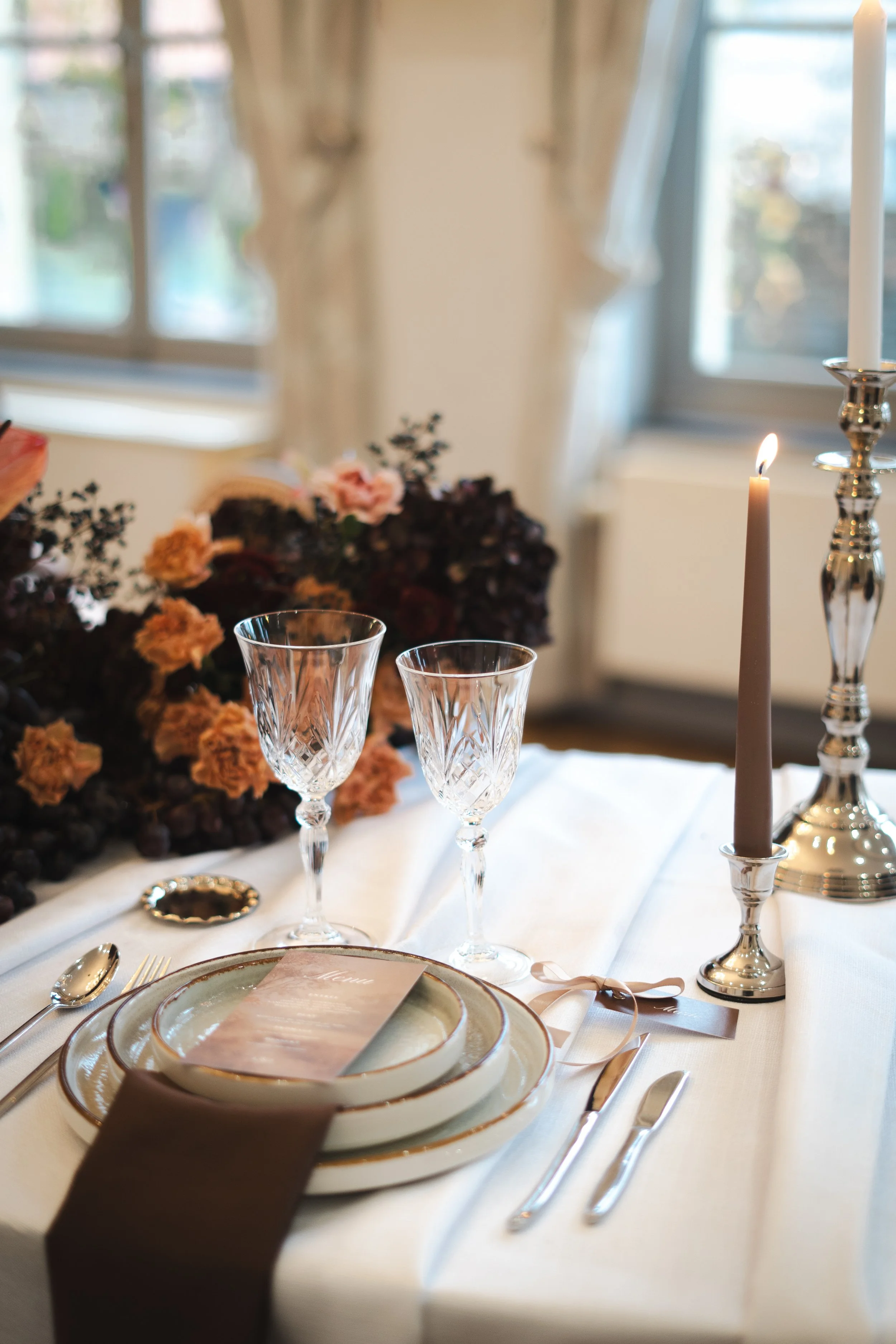 Une table élégante pour un repas, avec de la vaisselle en porcelaine, des verres à vin en cristal, des couverts en métal, deux chandelles allumées, un livre de menus, une nappe blanche, des fleurs séchées ou artificielles en arrière-plan, dans une pièce ensoleillée avec des fenêtres en arrière-plan.