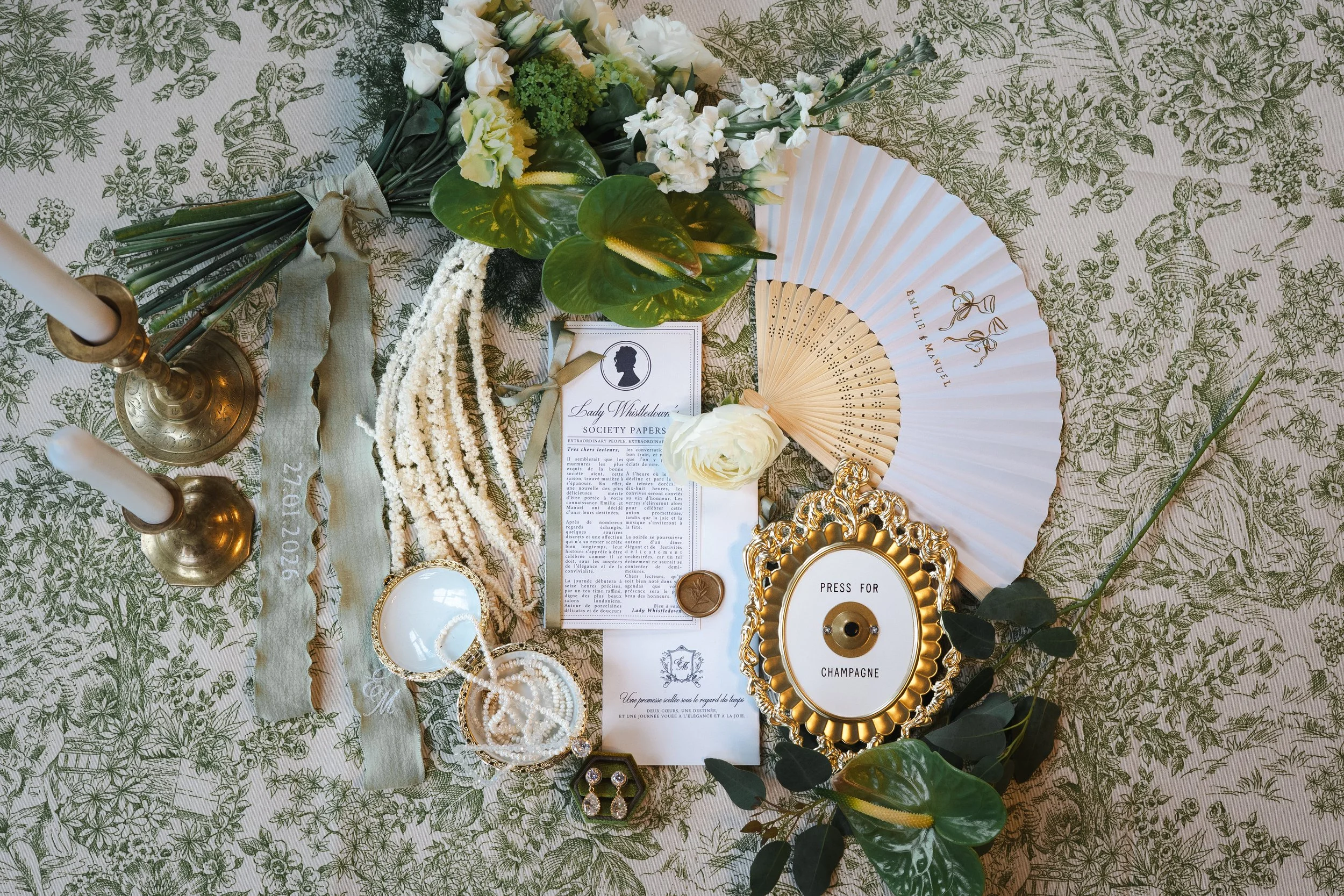 Disposition élégante avec des bougies, des fleurs, un éventail en bois, un presse-papiers doré, une carte, une pièce de monnaie, un disque avec un message et des feuilles vertes, le tout sur une nappe à motifs floraux.